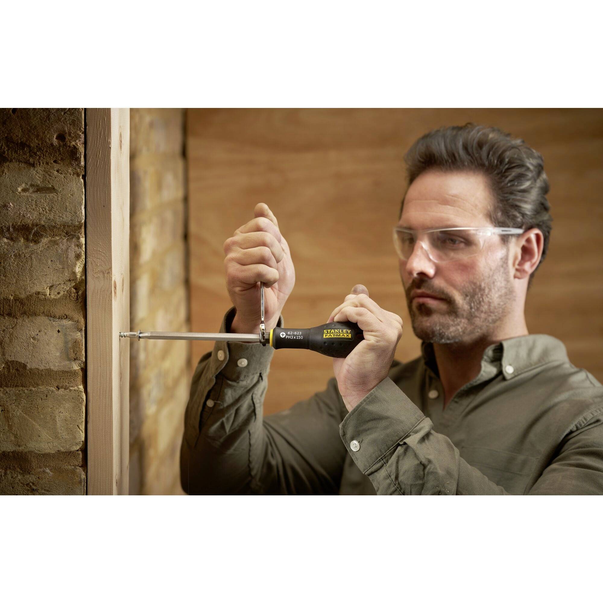 Osoba w okularach ochronnych używa śrubokręta, aby wkręcić śrubę w drewno. W tle widoczna jest ceglana ściana.