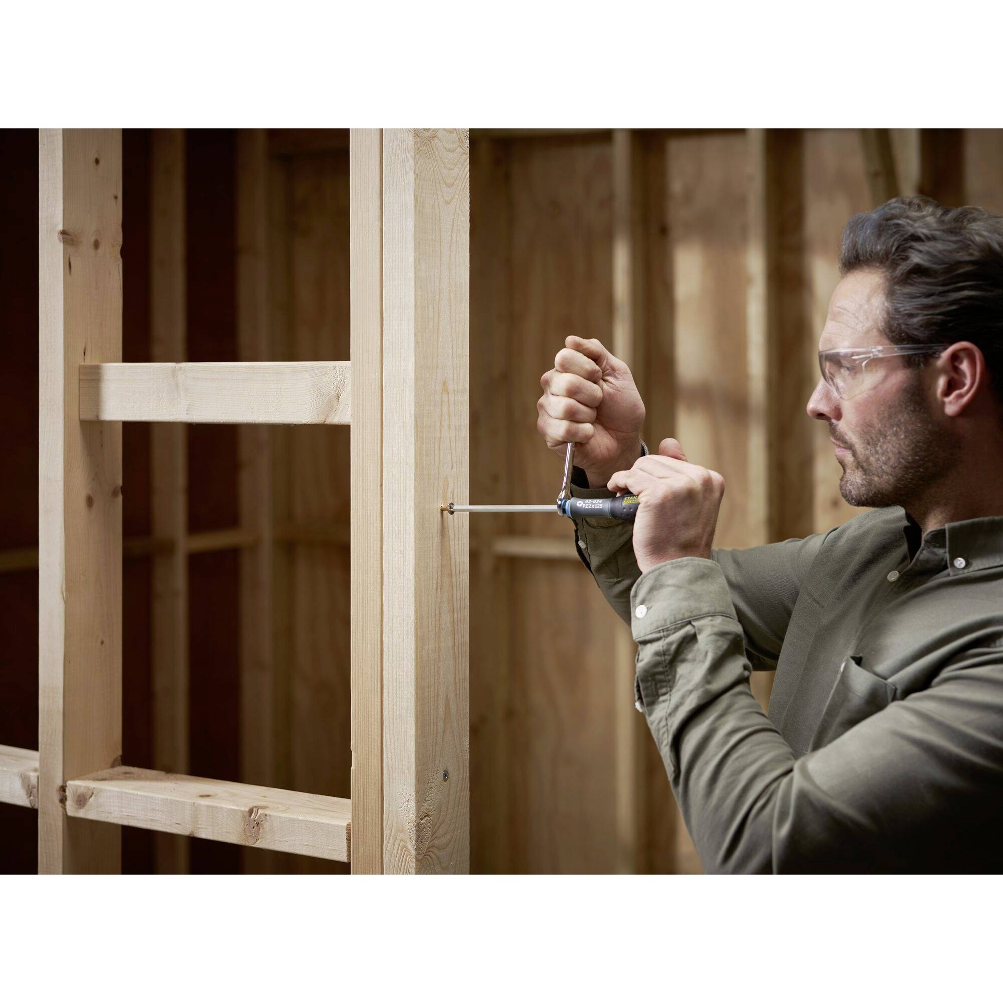 Mężczyzna w okularach ochronnych mocuje drewniane deski wkrętakiem w jakiejś konstrukcji. Może to być plac budowy lub remont.