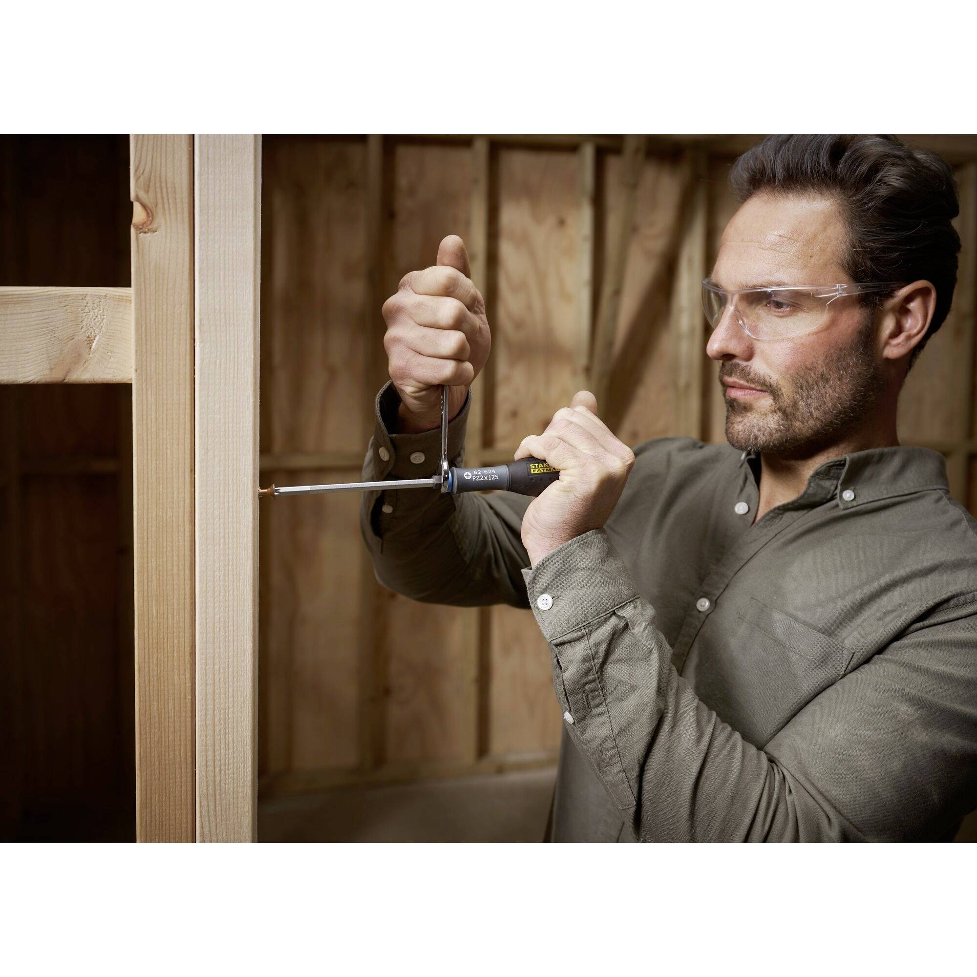 Osoba w okularach ochronnych montuje drewniane ramy wkrętakiem w niedokończonym pomieszczeniu.