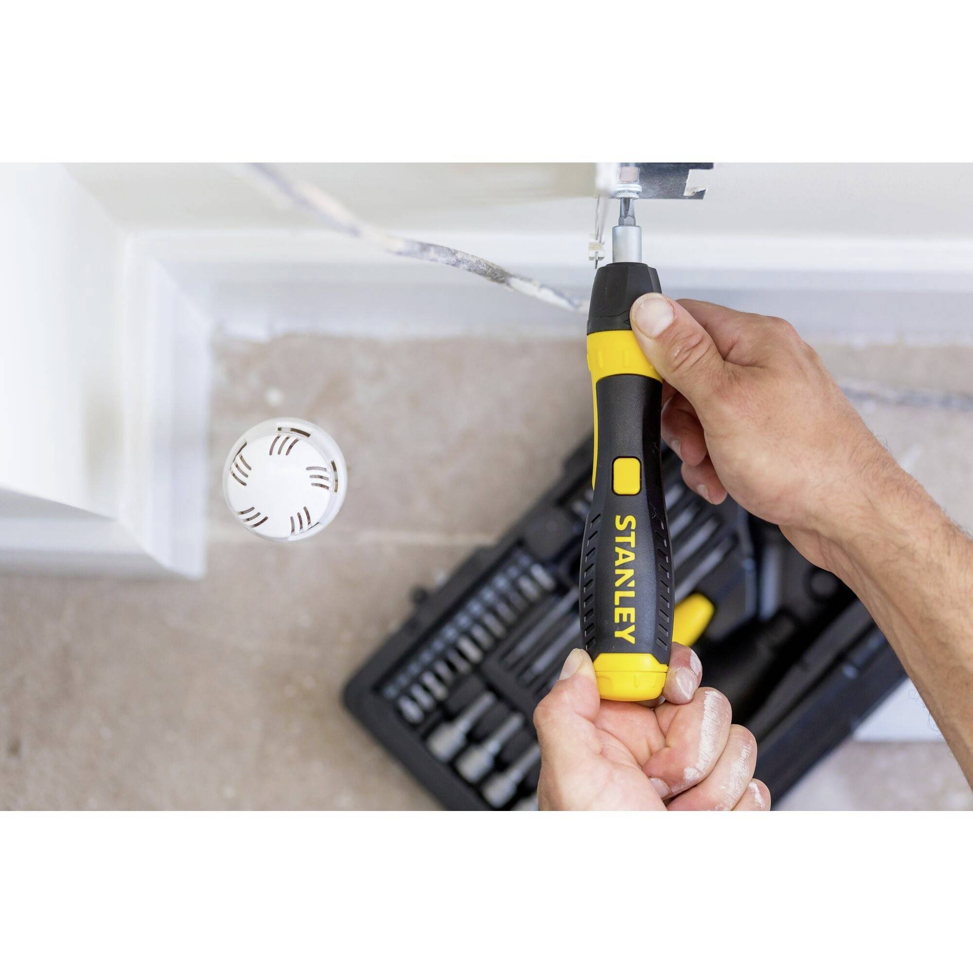 Osoba używa żółtego, elektrycznego wkrętaka, aby zamocować śrubę na białej ścianie. W tle widoczny jest zestaw narzędzi.