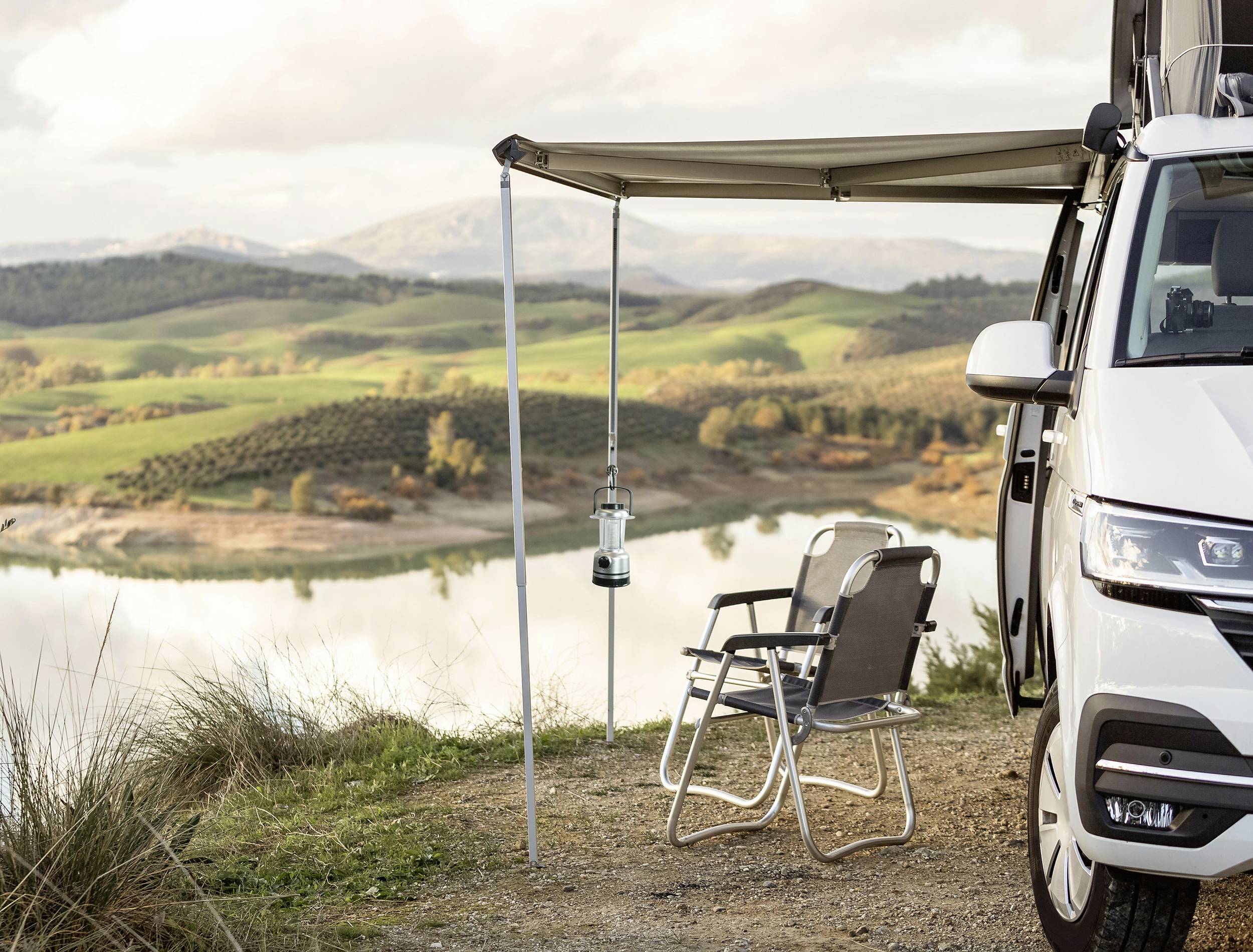 Kamper z markizą nad brzegiem jeziora. Dwa składane krzesła stoją pod markizą, w tle łagodne wzgórza i spokojne jezioro.