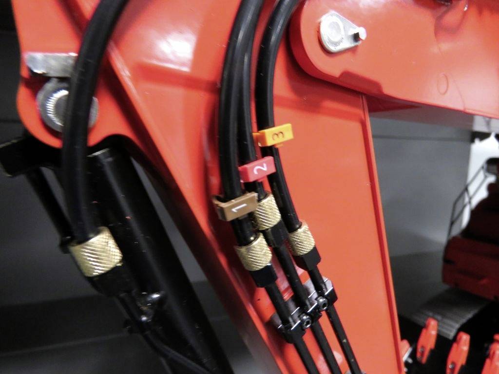 Zbliżenie hydraulicznych przewodów na czerwonej części maszyny budowlanej, oznaczonych kolorowymi plastikowymi klipsami wskazującymi pozycje przewodów.