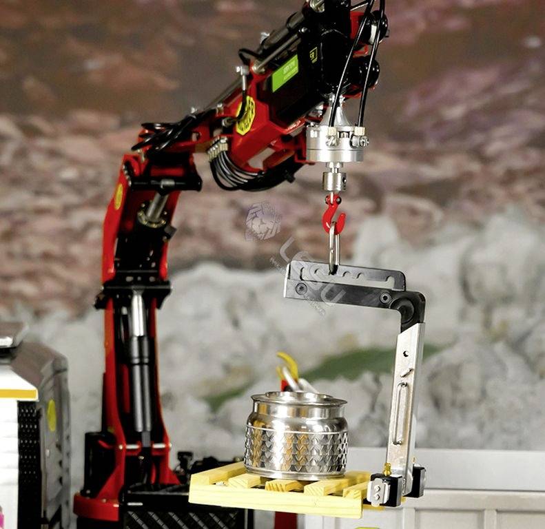 Czerwone ramię robotyczne podnosi srebrny pojemnik w przemysłowym otoczeniu. W tle widać rozmytą powierzchnię.