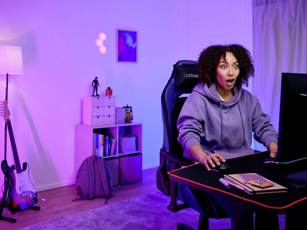 Osoba o kędzierzawych włosach siedzi zaskoczona przed komputerem w pokoju oświetlonym fioletowym światłem. W tle: lampa, regał, gitara elektryczna.