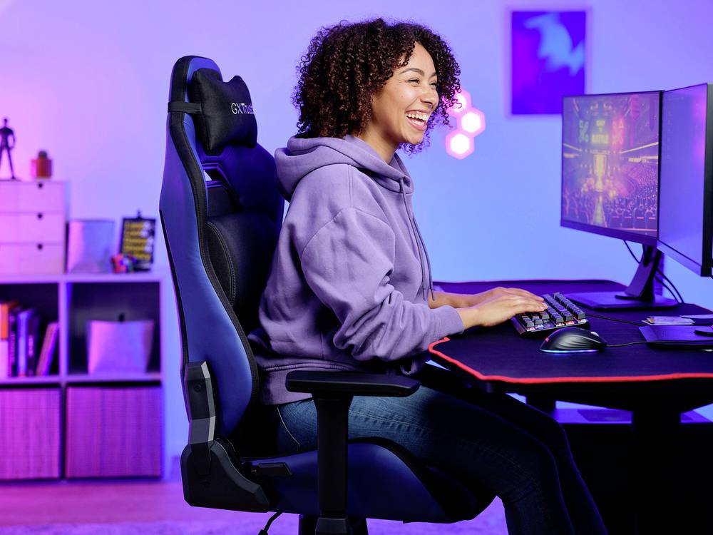 Osoba siedzi uśmiechnięta w fotelu gamingowym przed ekranem komputera. Pomieszczenie jest kolorowo oświetlone i urządzone w nowoczesnym stylu.