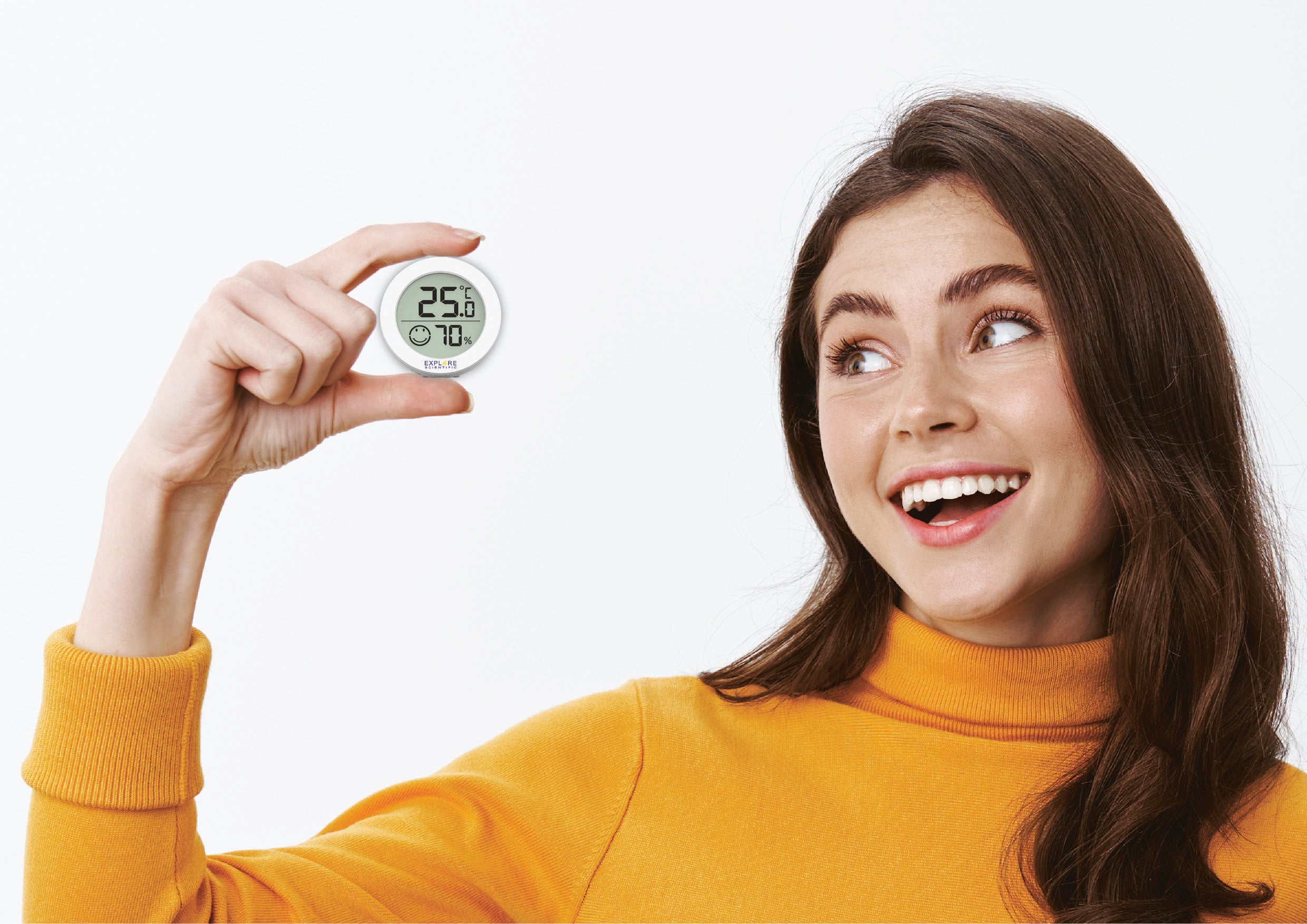 Kobieta w pomarańczowej bluzie trzyma okrągły cyfrowy termometr i higrometr, pokazujący 25,6°C i 70% wilgotności powietrza, uśmiechając się.