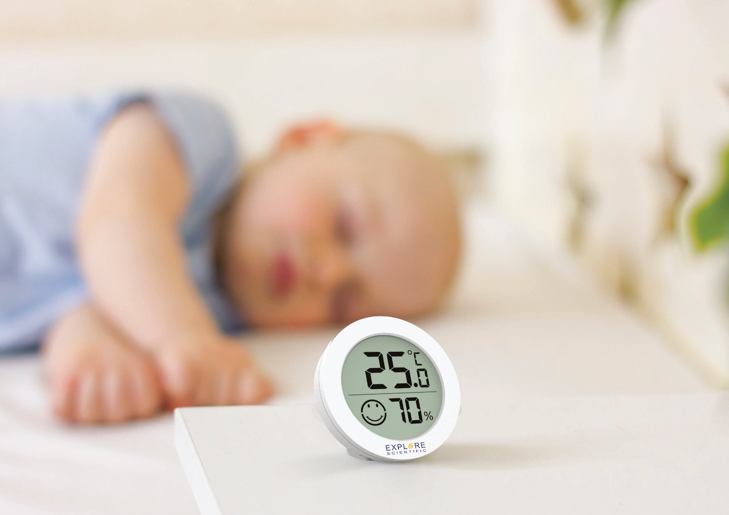 Śpiące niemowlę w tle; na pierwszym planie cyfrowy termo-higrometr wskazujący 25°C i 70% wilgotności powietrza.