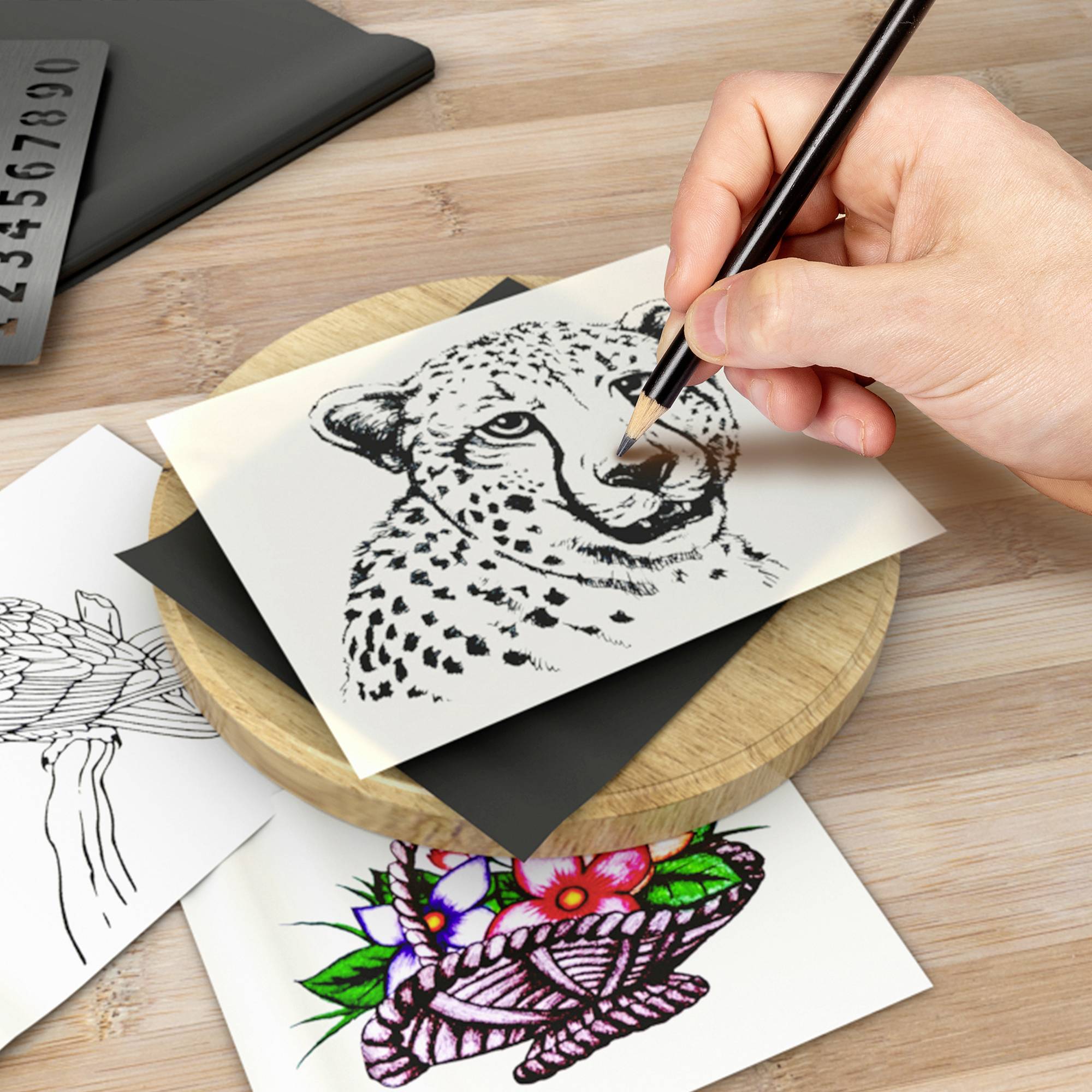 Jedna ręka rysuje głowę geparda na kartce papieru. Obok leżą kolejne ilustracje, w tym motywy kwiatowe.