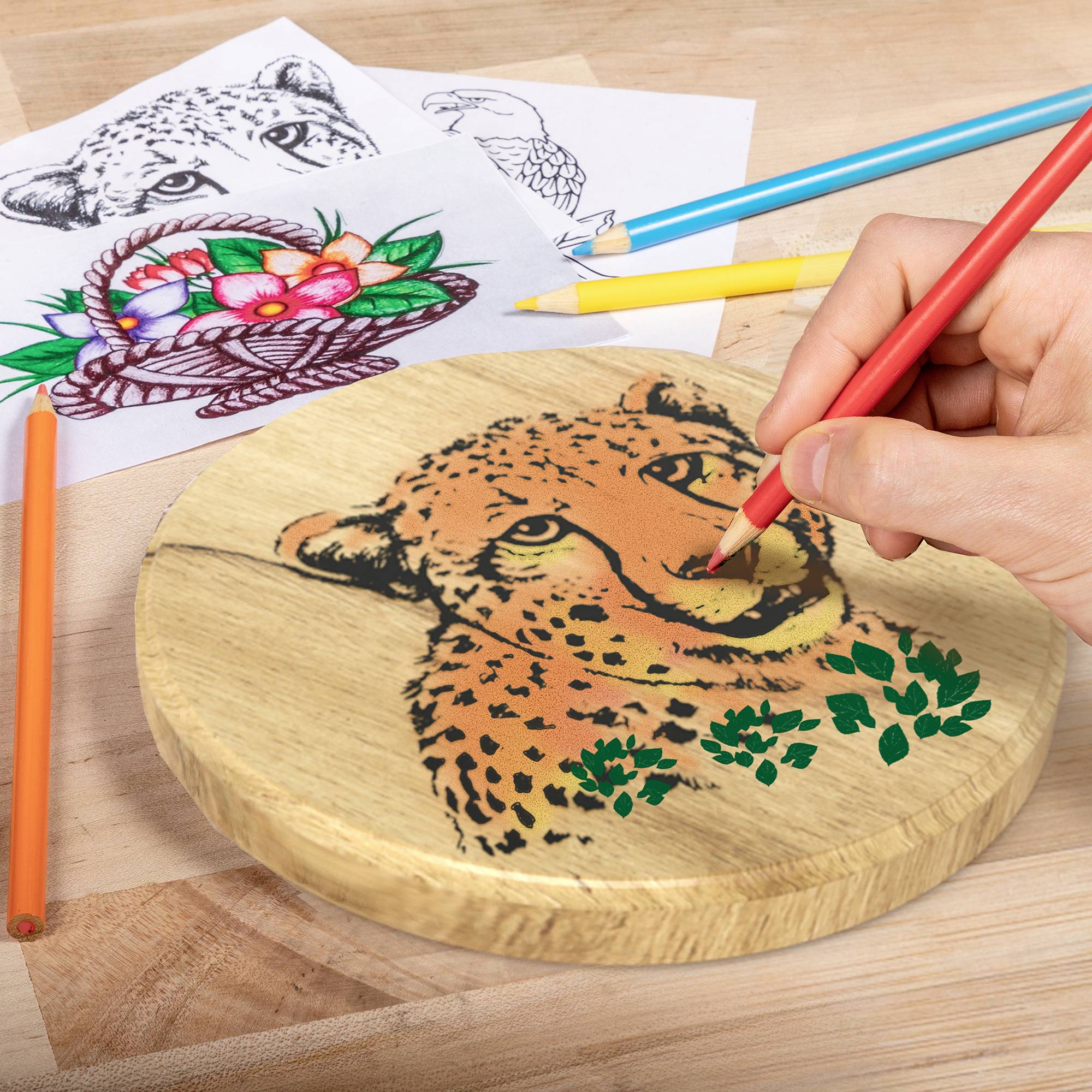 Osoba rysuje twarz geparda czerwonym piórem na drewnianej desce, otoczona kredkami i rysunkami zwierząt na papierze.