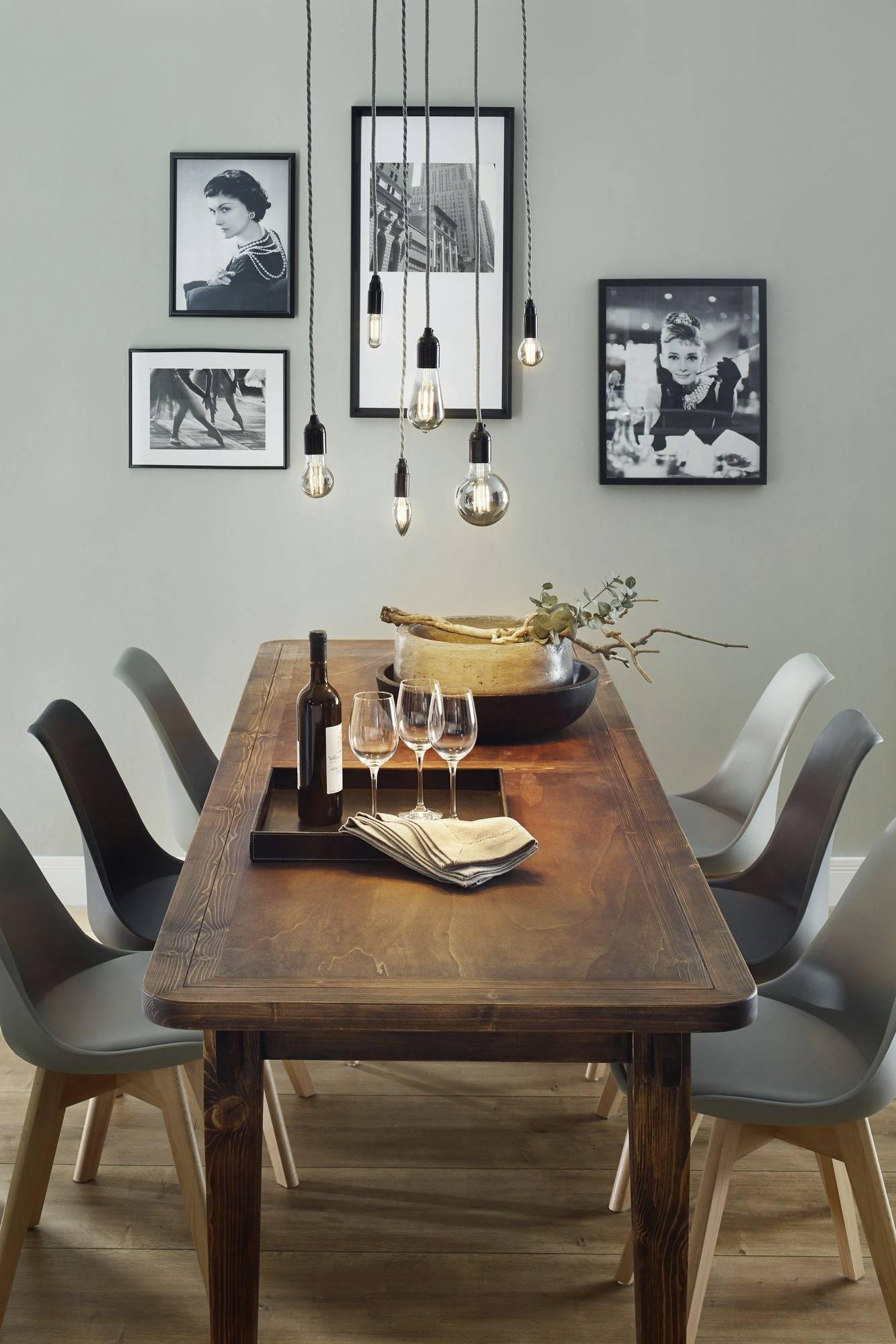 Jadalnia z drewnianym stołem, sześcioma szarymi krzesłami, wiszącymi żarówkami, winem i kieliszkami na tacy oraz czarno-białymi obrazami na ścianie.