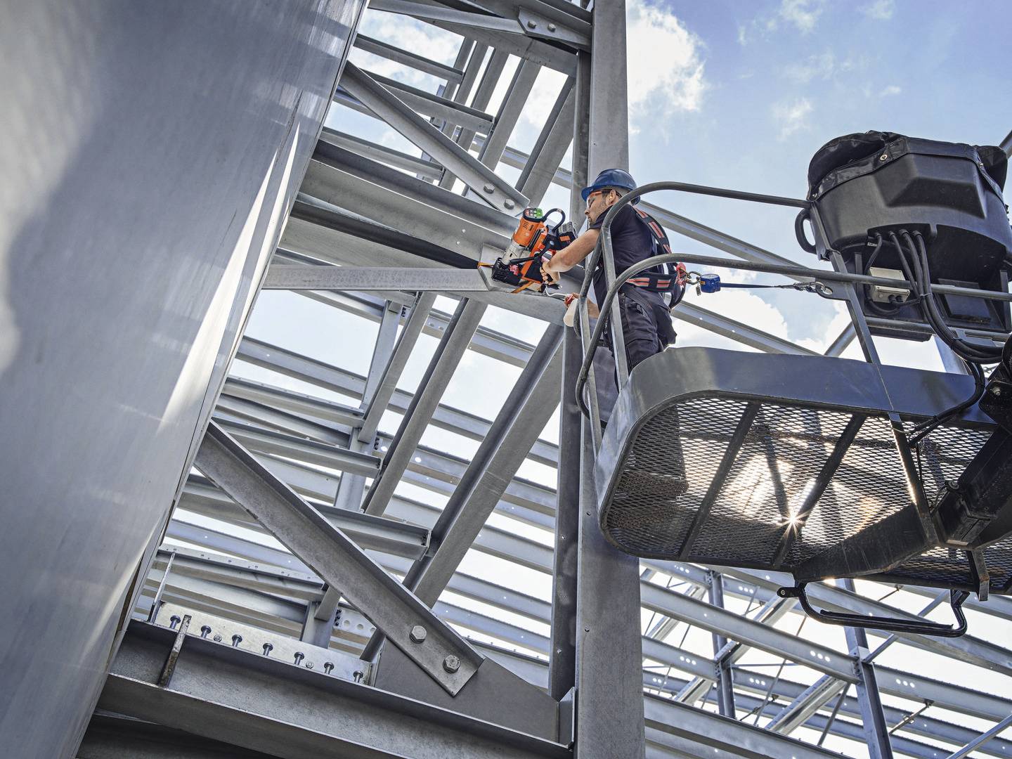Robotnik w odzieży ochronnej stoi na podnośniku i używa elektronarzędzia, pracując przy dużej stalowej konstrukcji.