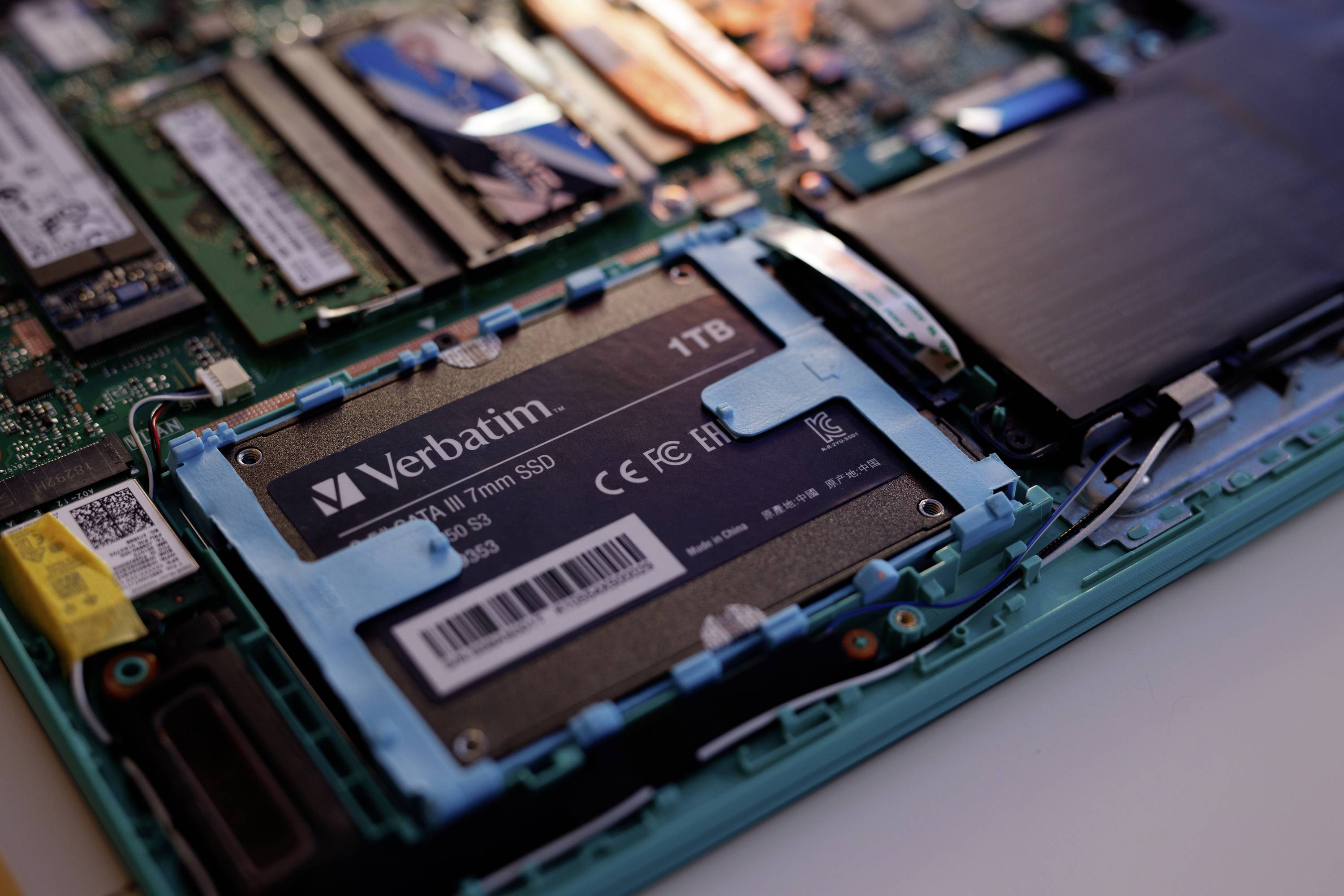 Zbliżenie otwartego laptopa z widocznym zainstalowanym dyskiem SSD Verbatim o pojemności 1 TB, otoczonym elementami sprzętu komputerowego.