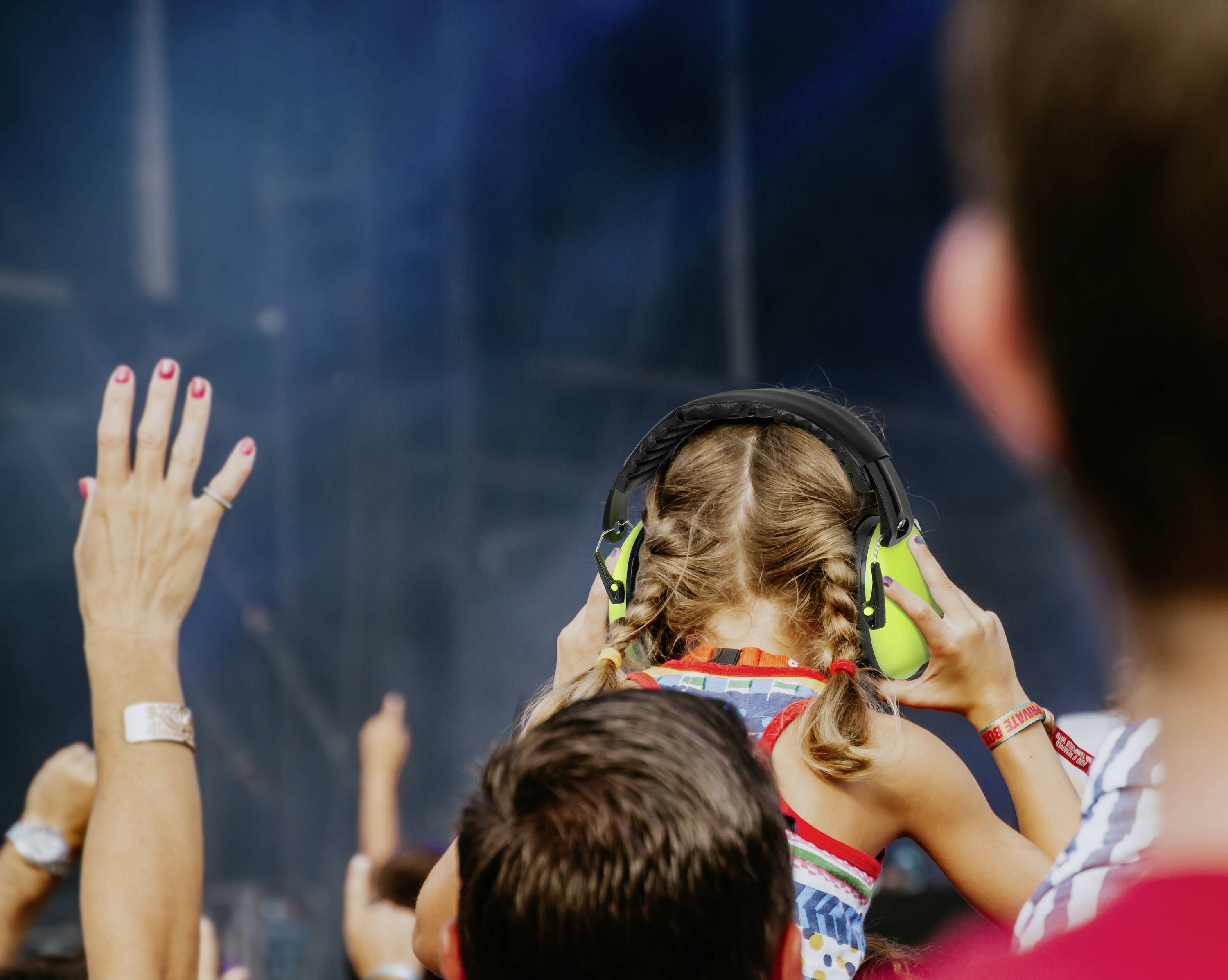 Dziecko w ochronnikach słuchu siedzi na ramionach dorosłego i słucha koncertu na żywo, podczas gdy publiczność podnosi ręce.