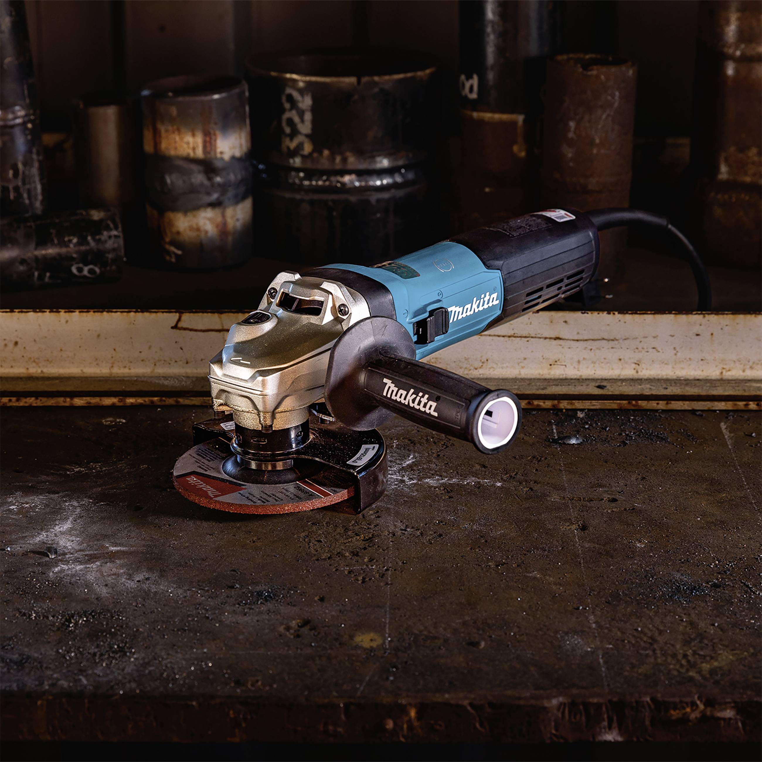 Szlifierka kątowa leży na stole warsztatowym, otoczona metalowymi elementami. Jest głównie niebieska, z czarną rękojeścią i czerwoną tarczą ścierną.