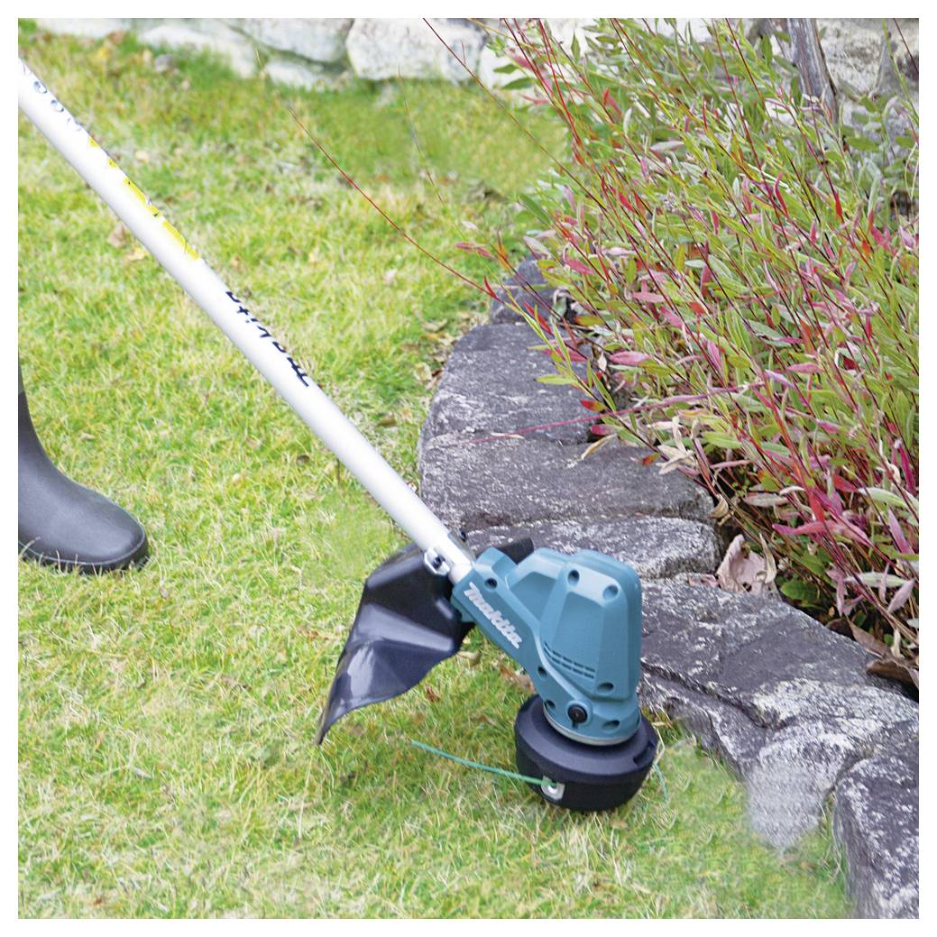 Osoba korzystająca z zasilanej baterią przycinaczki do trawy do cięcia trawy obok grządki ogrodowej z roślinami, nosząca czarne buty dla ochrony.