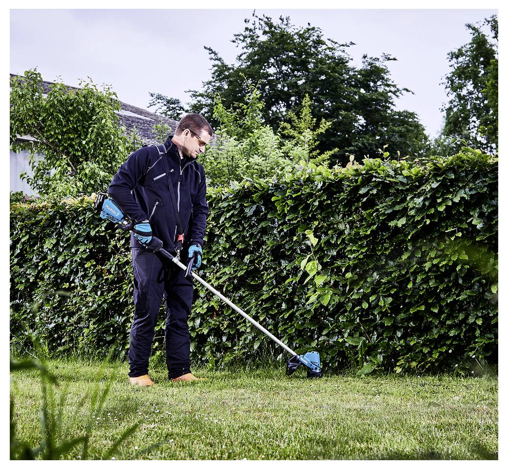Osoba nosząca rękawiczki i odzież ochronną przystrzyga żywopłot za pomocą elektrycznej nożycy do żywopłotu w ogrodzie z gęstą zielenią.