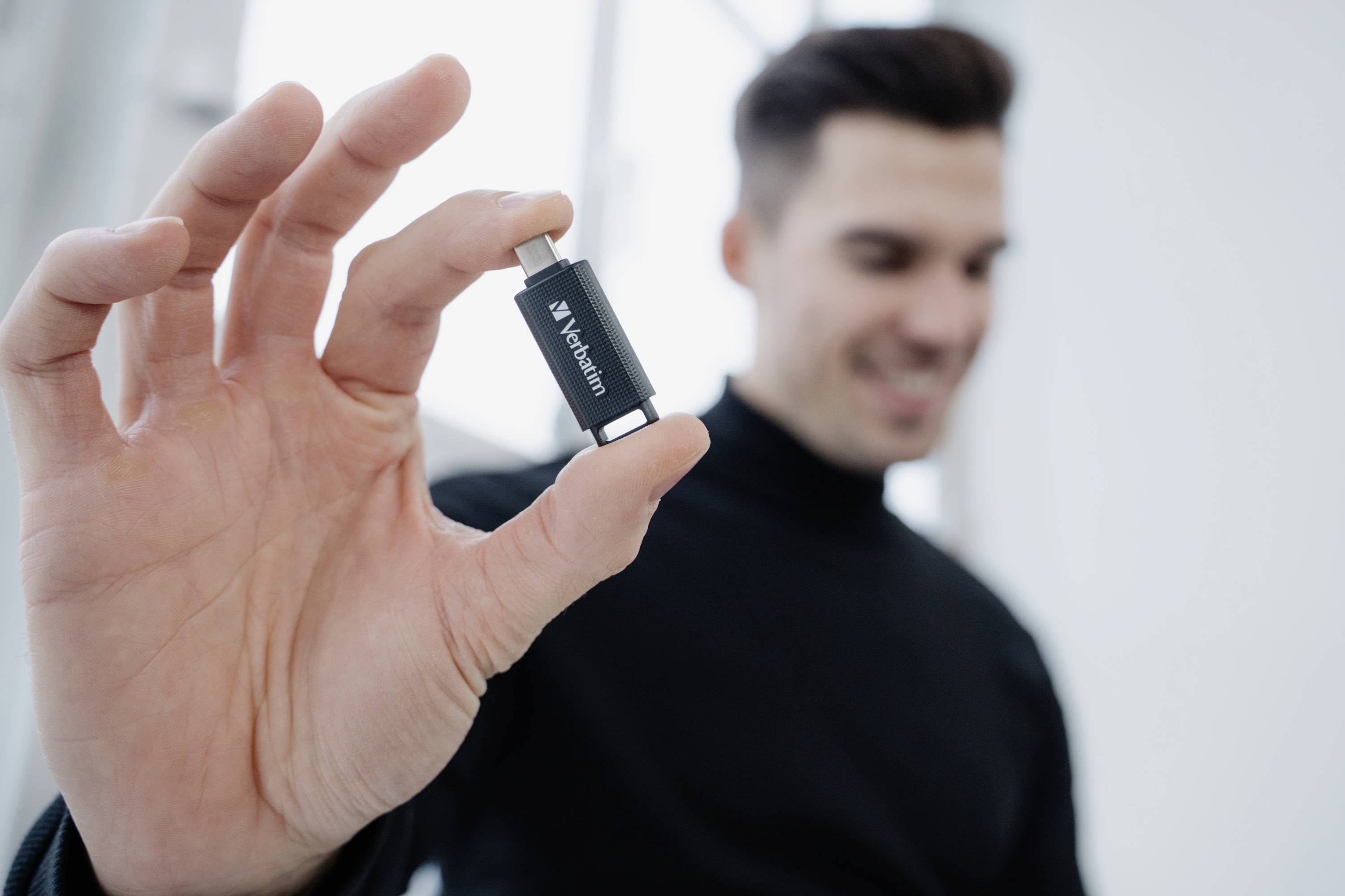 Osoba trzyma mały pendrive w ręku, uśmiechając się na rozmytym, jasno oświetlonym tle.