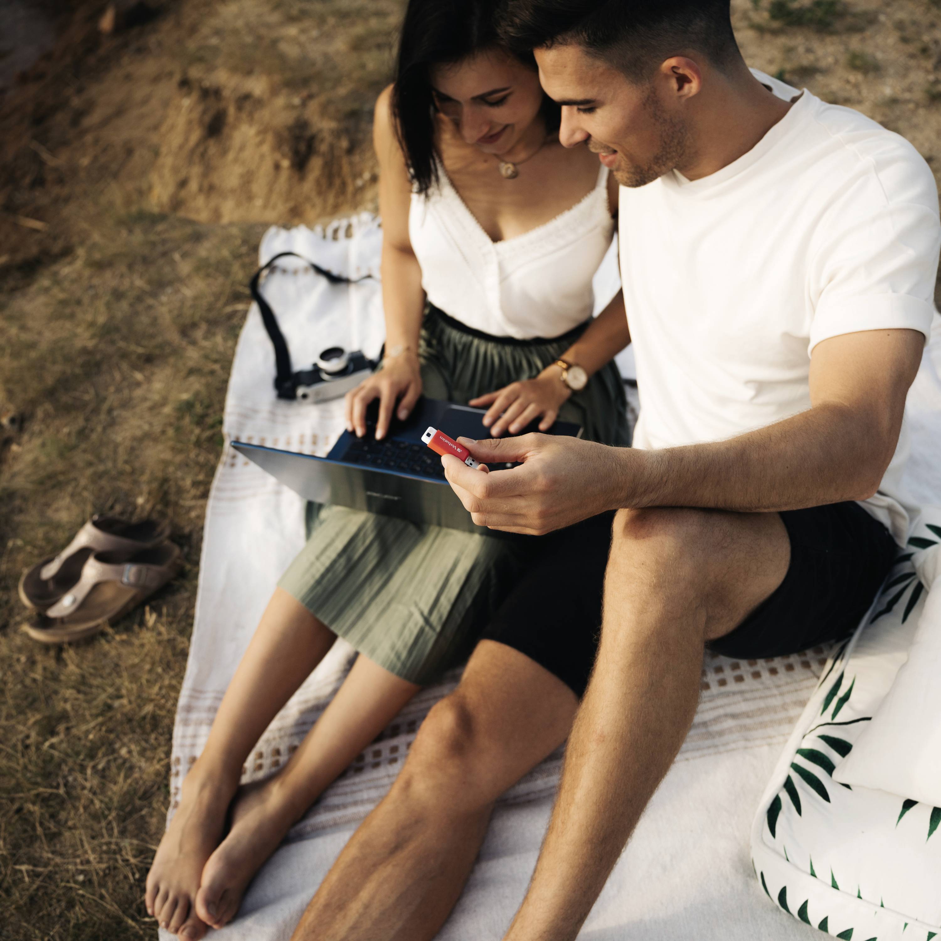 Para siedzi na kocu na świeżym powietrzu i wspólnie przygląda się tabletowi. Śmieją się i wyglądają na zrelaksowanych.