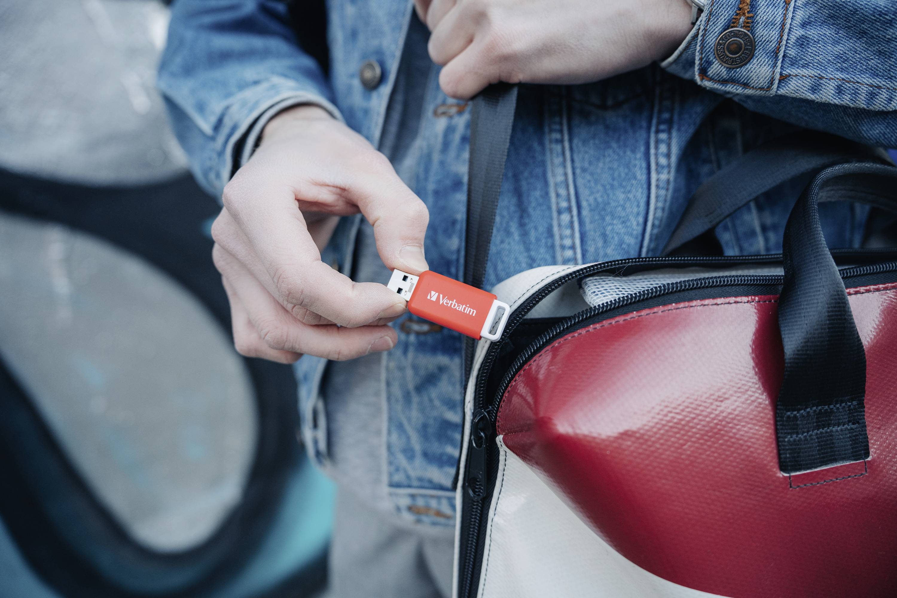 Osoba wkłada czerwoną pamięć USB do torby. Ma na sobie jeansową kurtkę. Pendrive ma nadruk 'Verbatim'.
