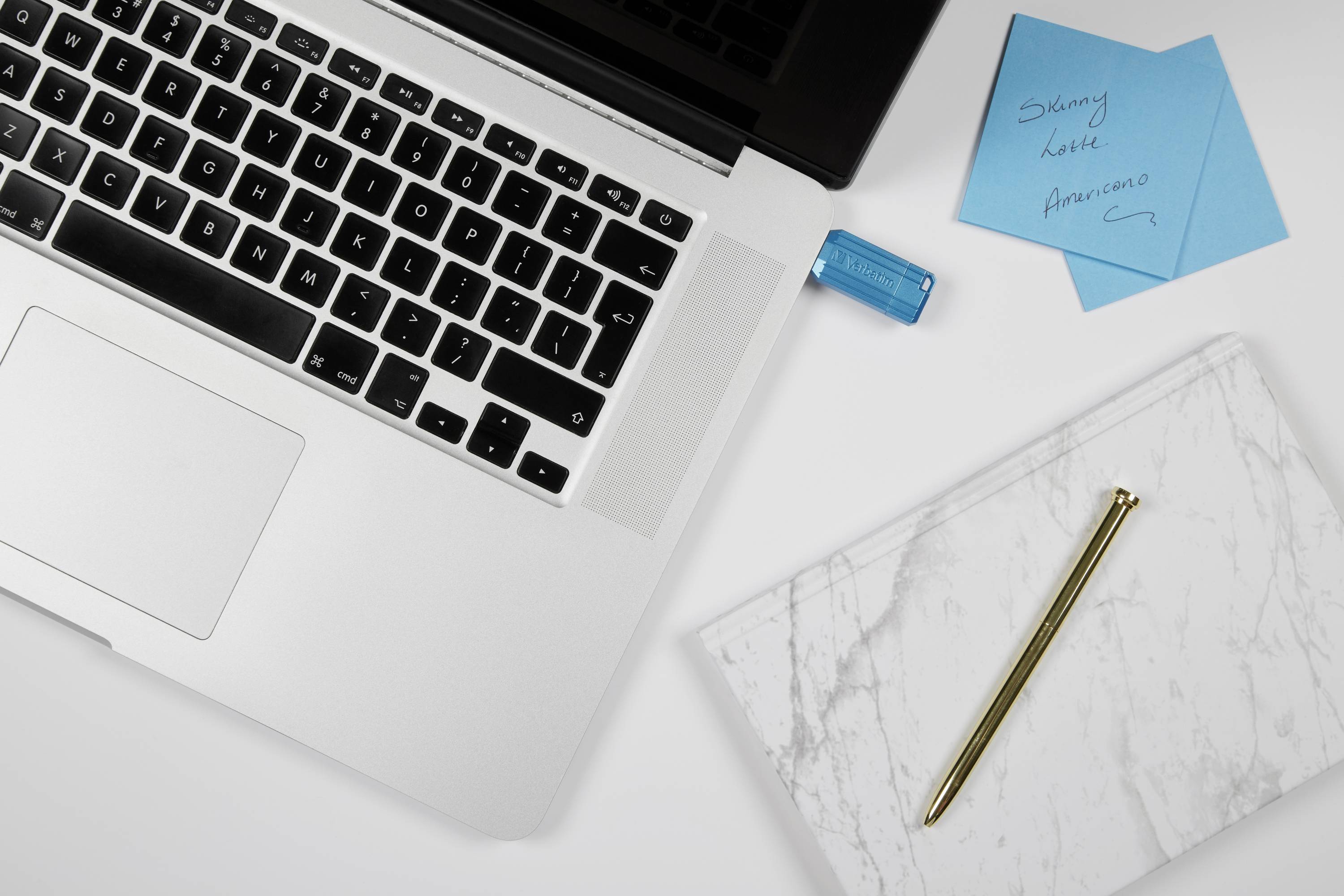 Laptop na biurku obok notes'a z marmurowej okładki, złotego pióra, niebieskich karteczek samoprzylepnych i pendrive'a.