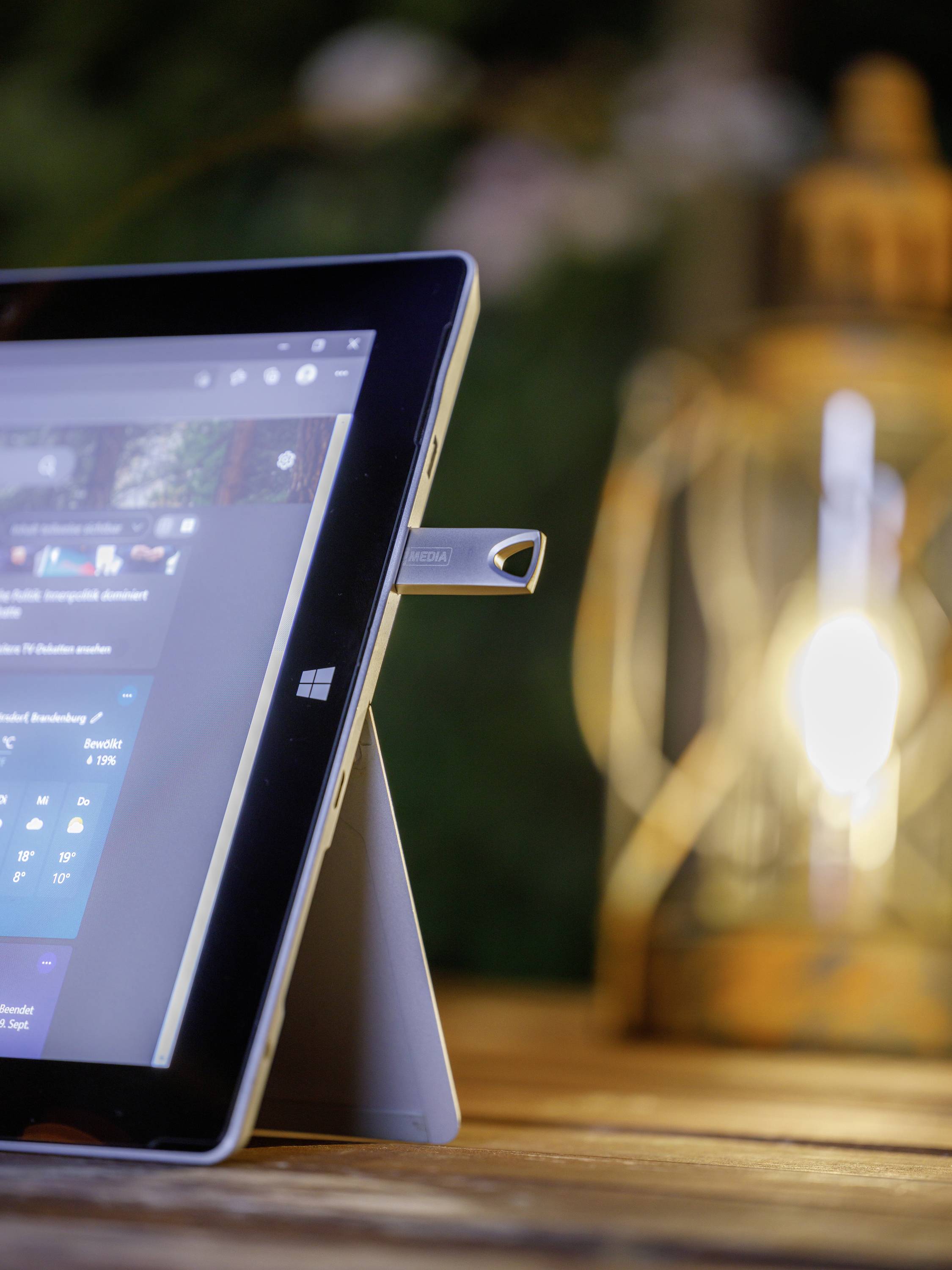 Tablet z podłączonym pendrive'em stoi na stole obok ciepło świecącej lampy, w otoczeniu rozmytego tła.