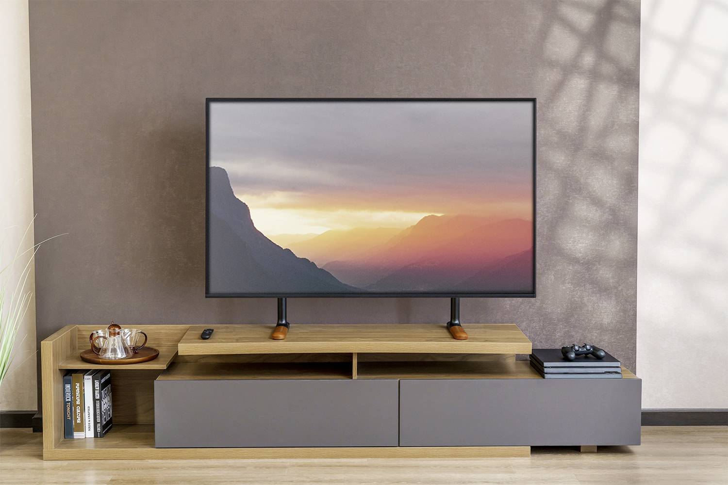 Płaski telewizor na drewnianej szafce przed brązową ścianą, wyświetlający zachód słońca w górskim krajobrazie. Po lewej stronie taca z filiżankami.