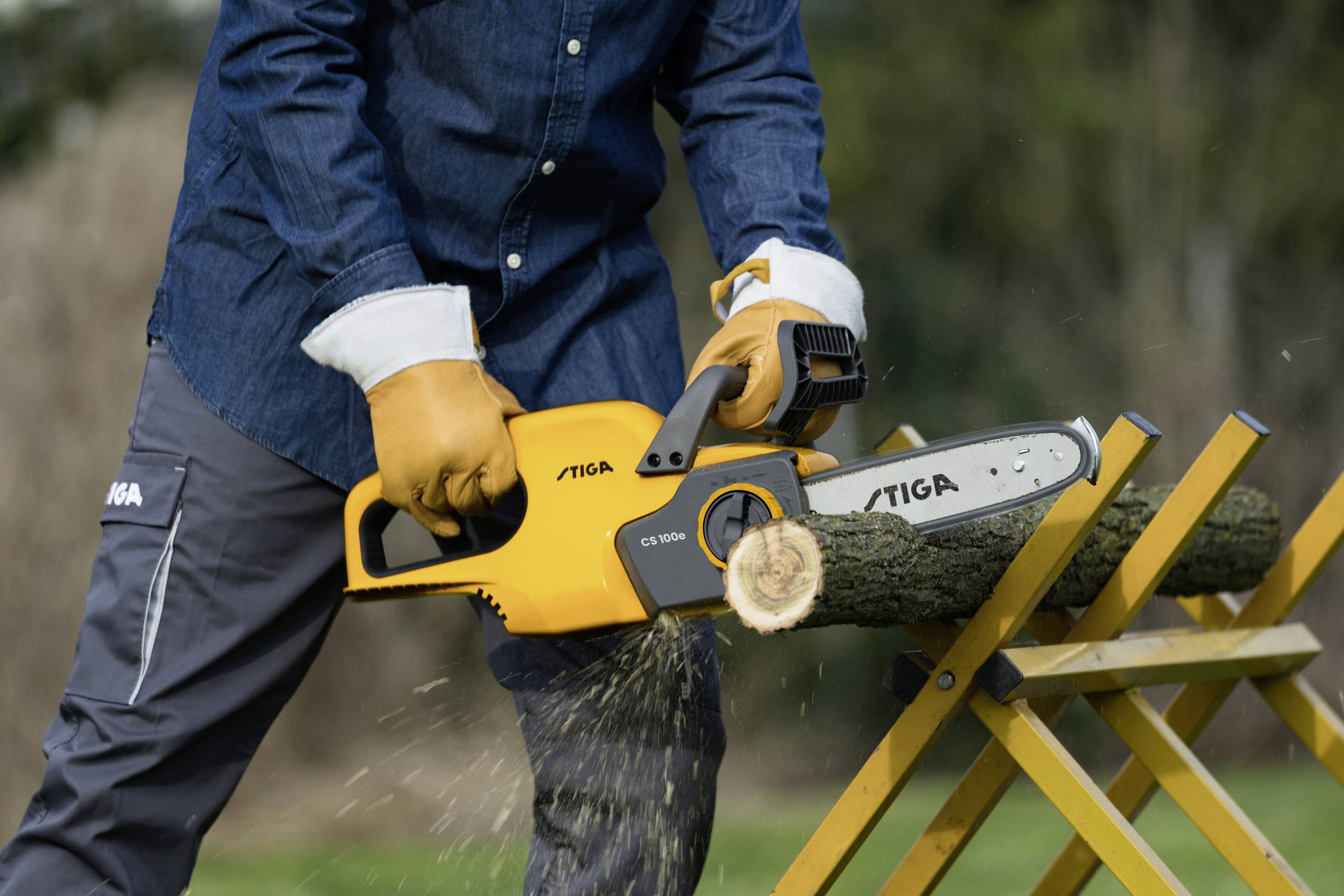 Osoba tnie piłą łańcuchową elektryczną drewniany pień, który leży na koźle stolarskim. Nosi przy tym odzież ochronną.