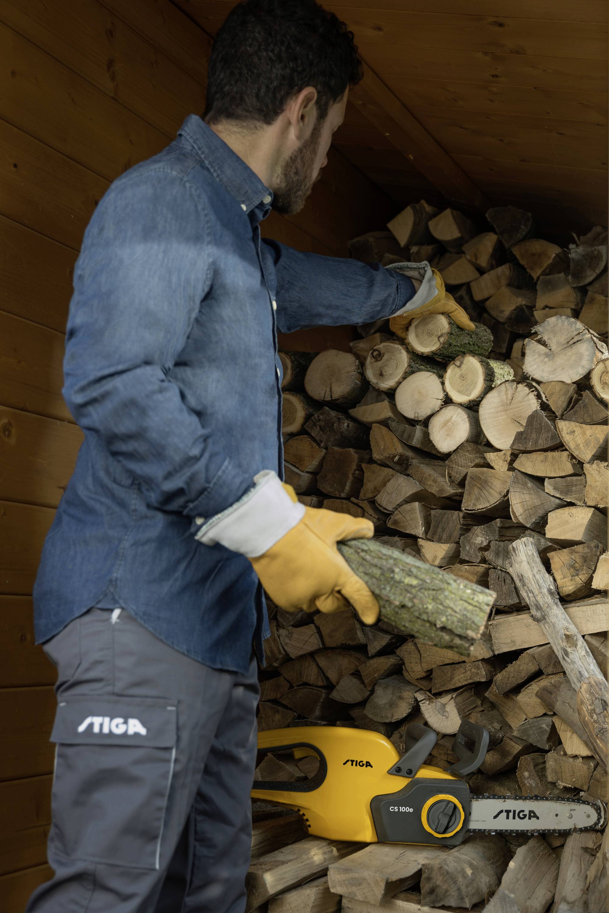 Mężczyzna w ubraniu roboczym układa drewno opałowe w szopie. Obok niego na podłodze leży piła motorowa.