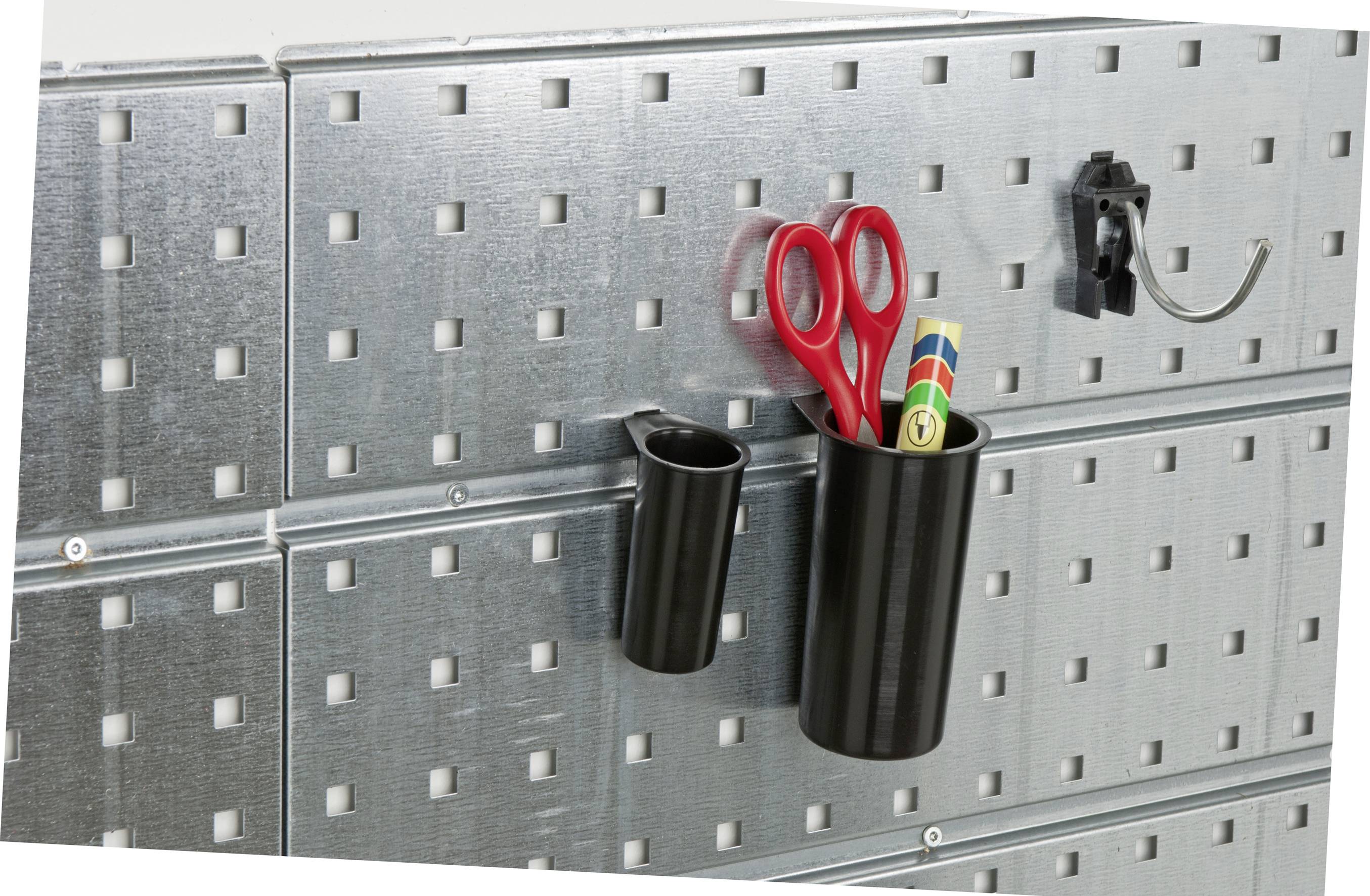 Metalowa ściana z kwadratowymi wycięciami, na której wiszą dwa czarne pojemniki; jeden z nich trzyma czerwone nożyczki i przybory malarskie.