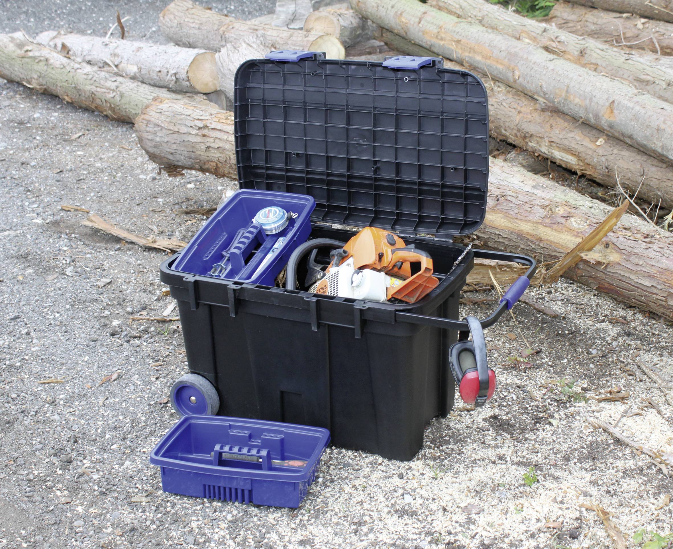 Otwarta skrzynka narzędziowa z piłą łańcuchową i innymi narzędziami stoi przed ułożonymi w stos pniami drzew na żwirowym podłożu.