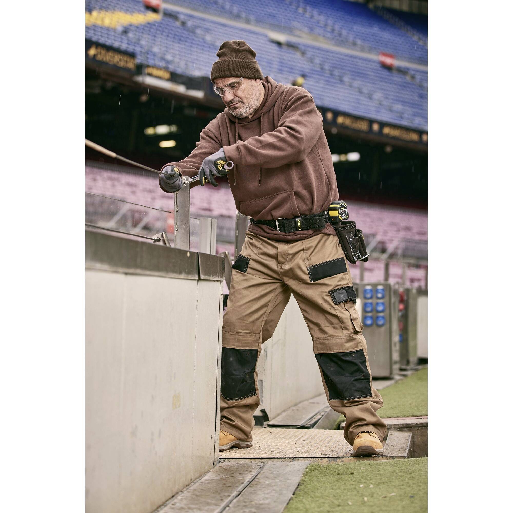 Robotnik budowlany w stroju ochronnym pracuje z narzędziami przy metalowej barierze na dużym stadionie. Pada lekki deszcz.