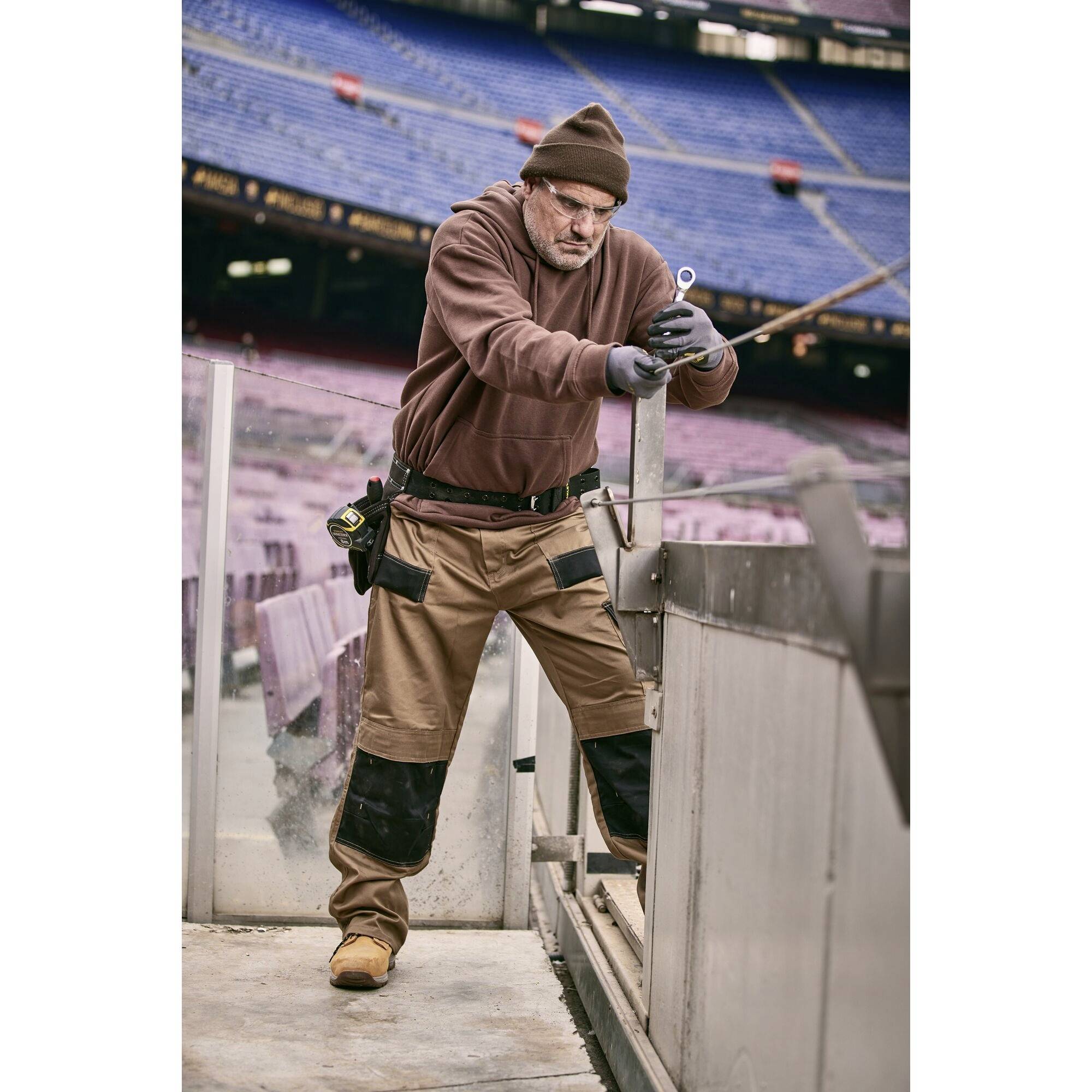 Osoba w stroju roboczym naprawia metalową barierę na pustym stadionie. Otoczenie ukazuje puste trybuny i rzędy siedzeń.