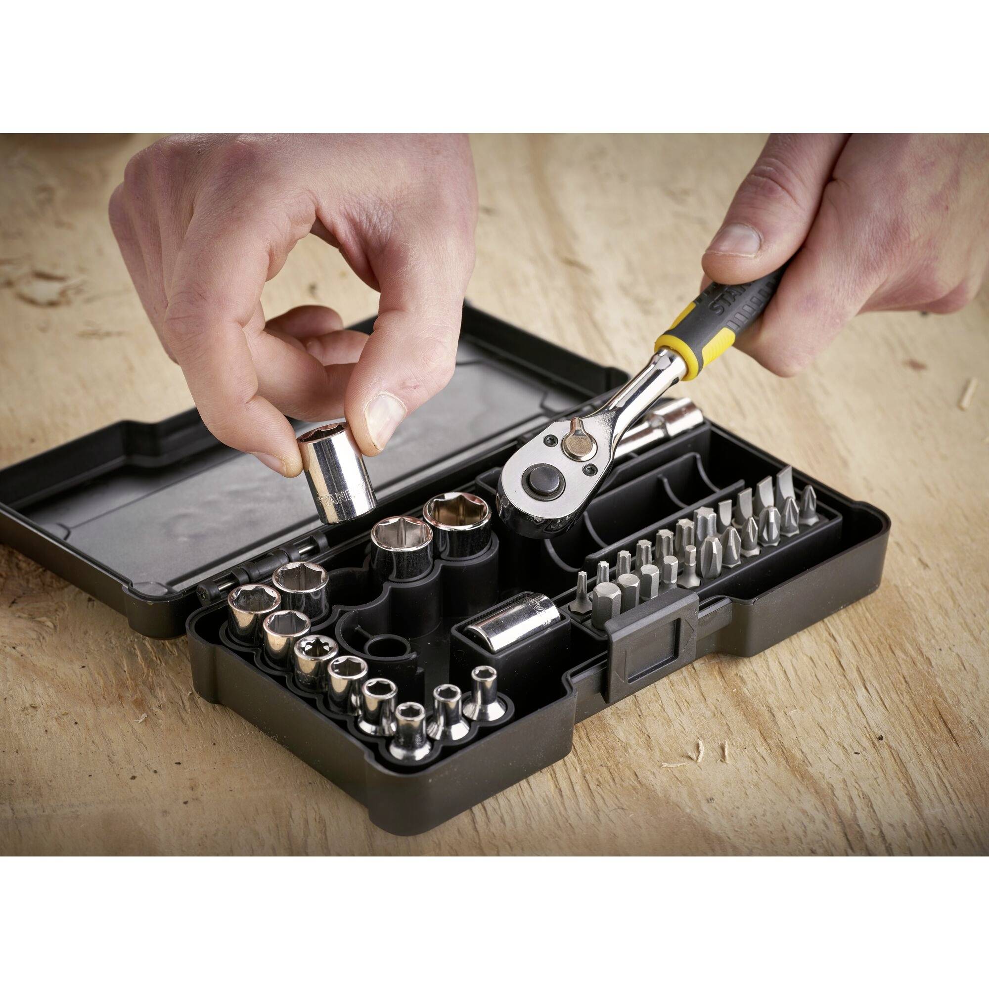 Zestaw kluczy nasadowych w otwartym czarnym pudełku. Jedna ręka trzyma klucz nasadowy, druga - grzechotkę na stole warsztatowym.