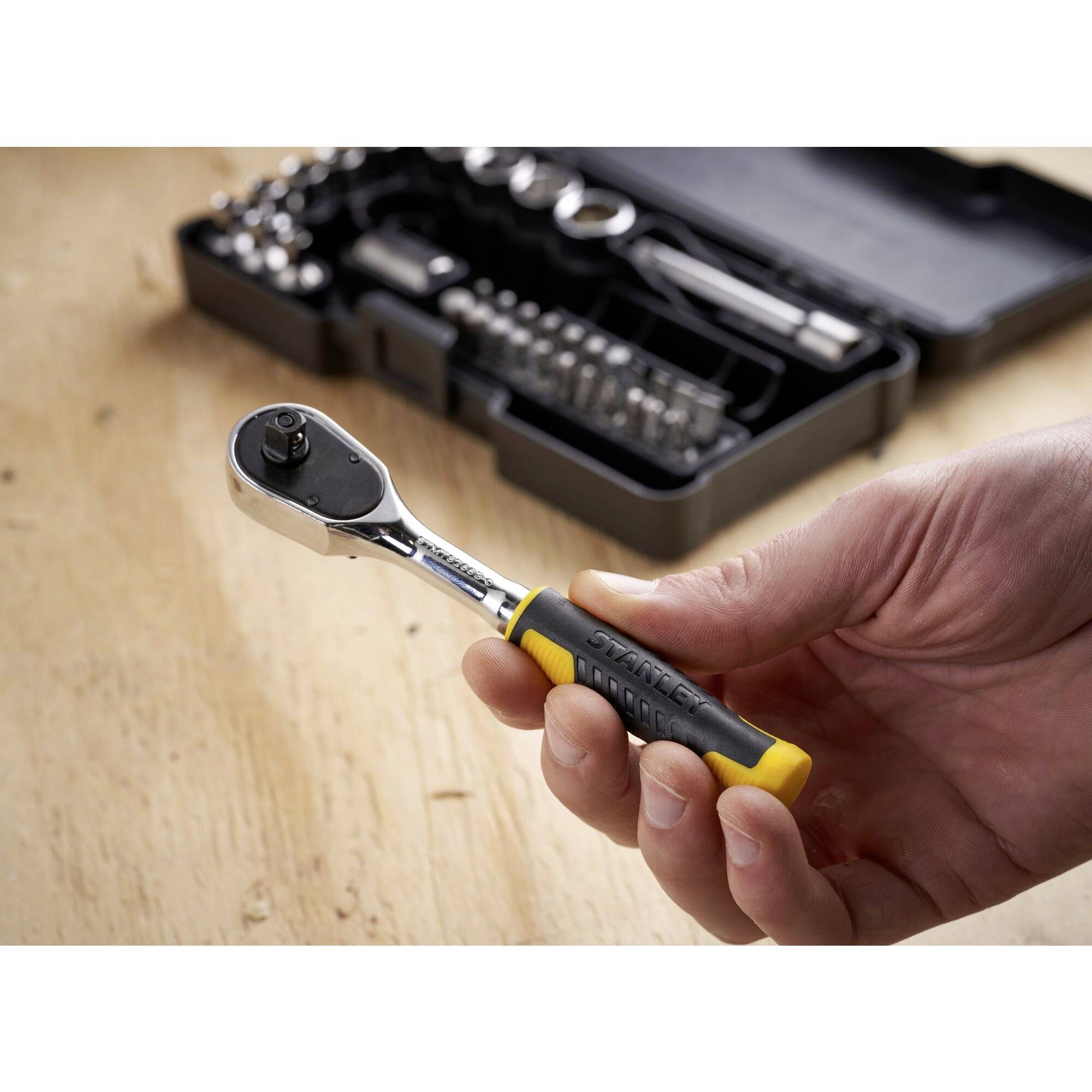 Dłoń trzyma grzechotkę przed otwartą skrzynką narzędziową z różnymi nasadkami i bitami na drewnianym stole.