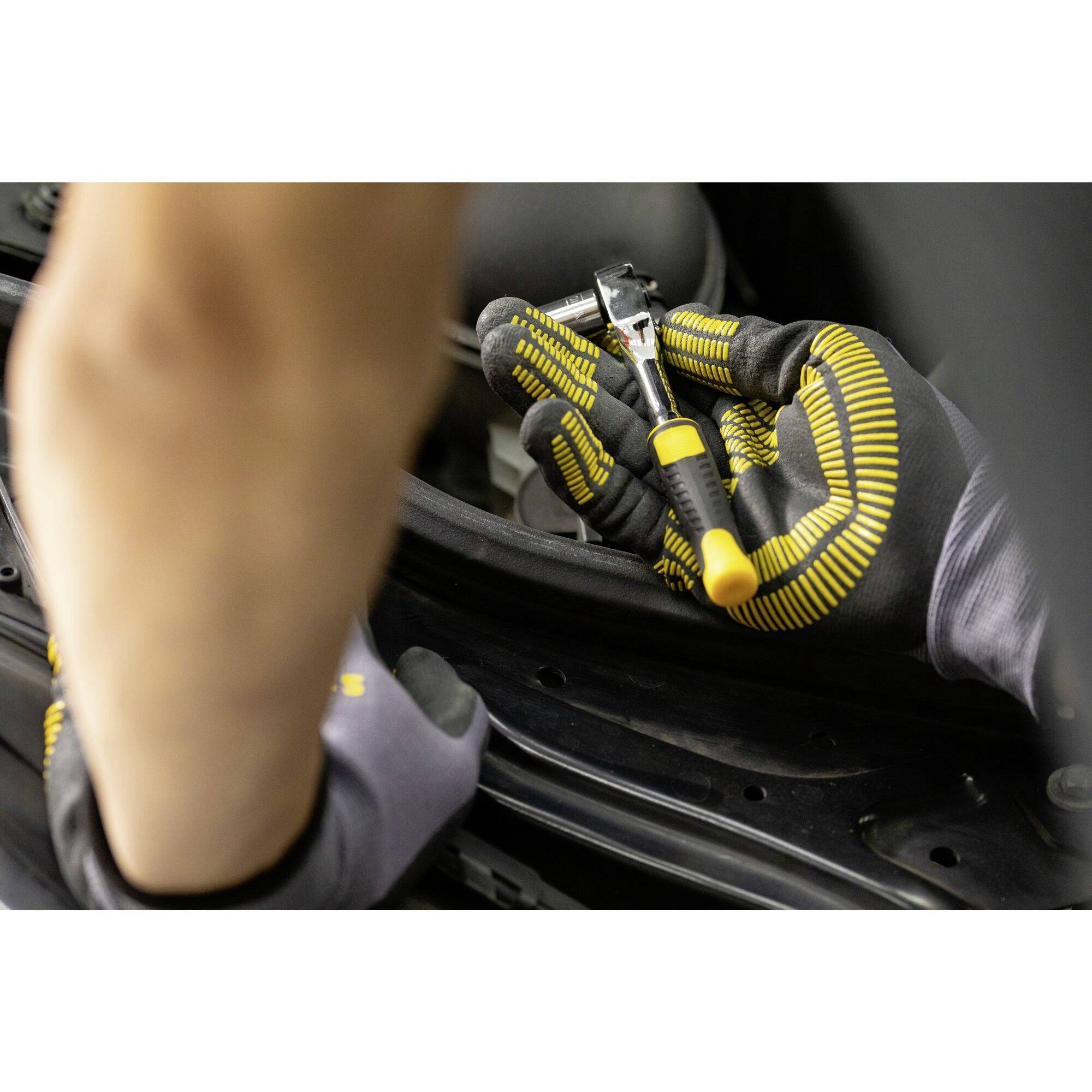 Osoba w czarnych, żółto prążkowanych rękawiczkach naprawia samochód kluczem płaskim pod otwartą maską silnika.