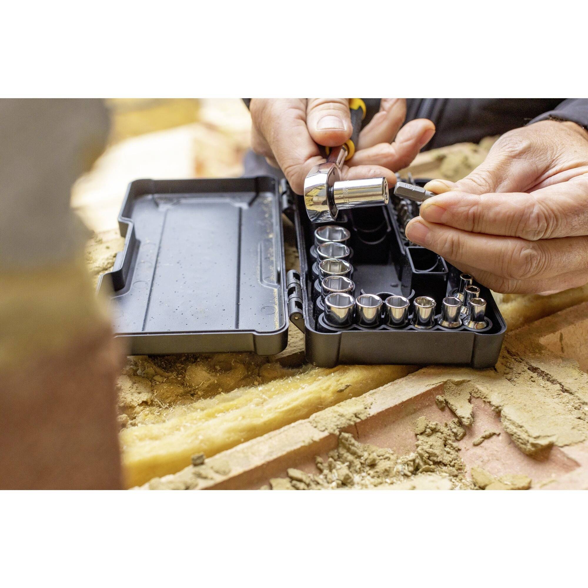 Dwie ręce pracują z zestawem kluczy nasadowych. Jedna ręka trzyma grzechotkę, druga nakładkę nasadową. Narzędzia leżą w otwartej skrzynce.