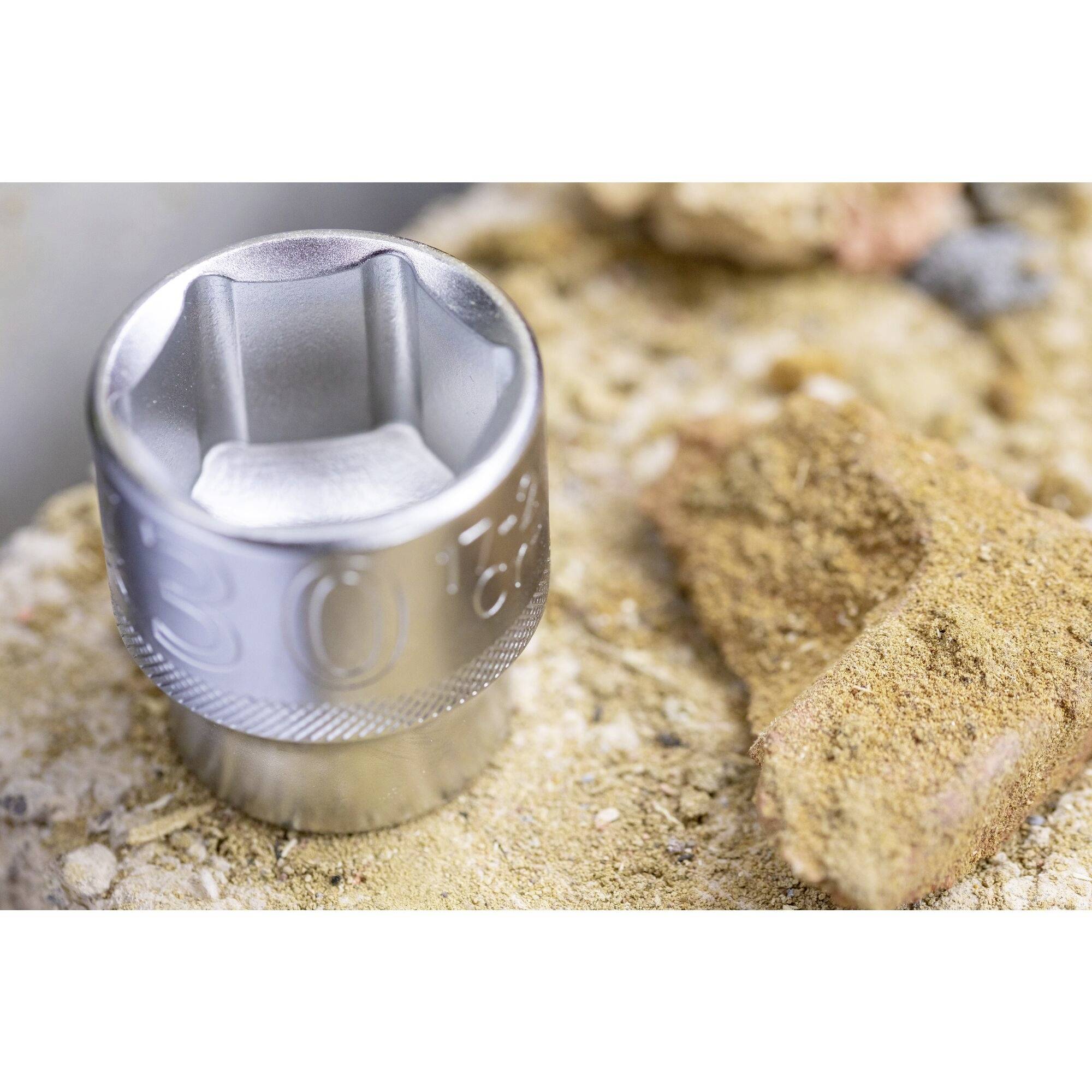 Zbliżenie srebrnego klucza nasadowego na piaszczystym podłożu, podkreślające szorstkość i zużycie narzędzia.