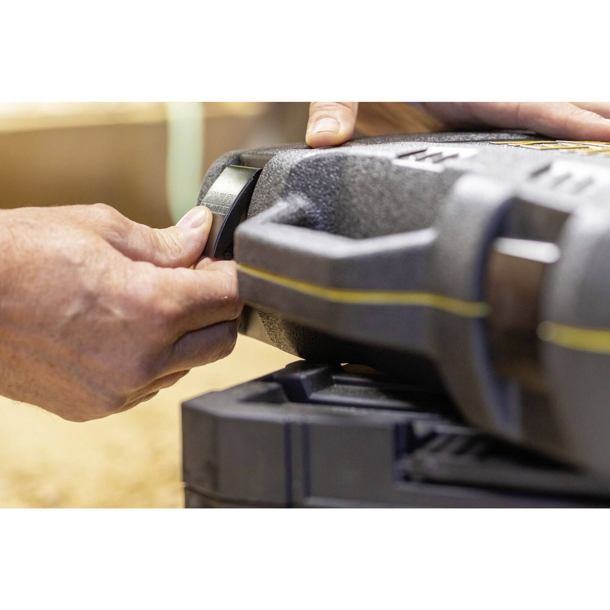 Zbliżenie dłoni otwierającej zamek ciężkiej czarnej walizki z tworzywa sztucznego. Walizka posiada żółte akcenty.