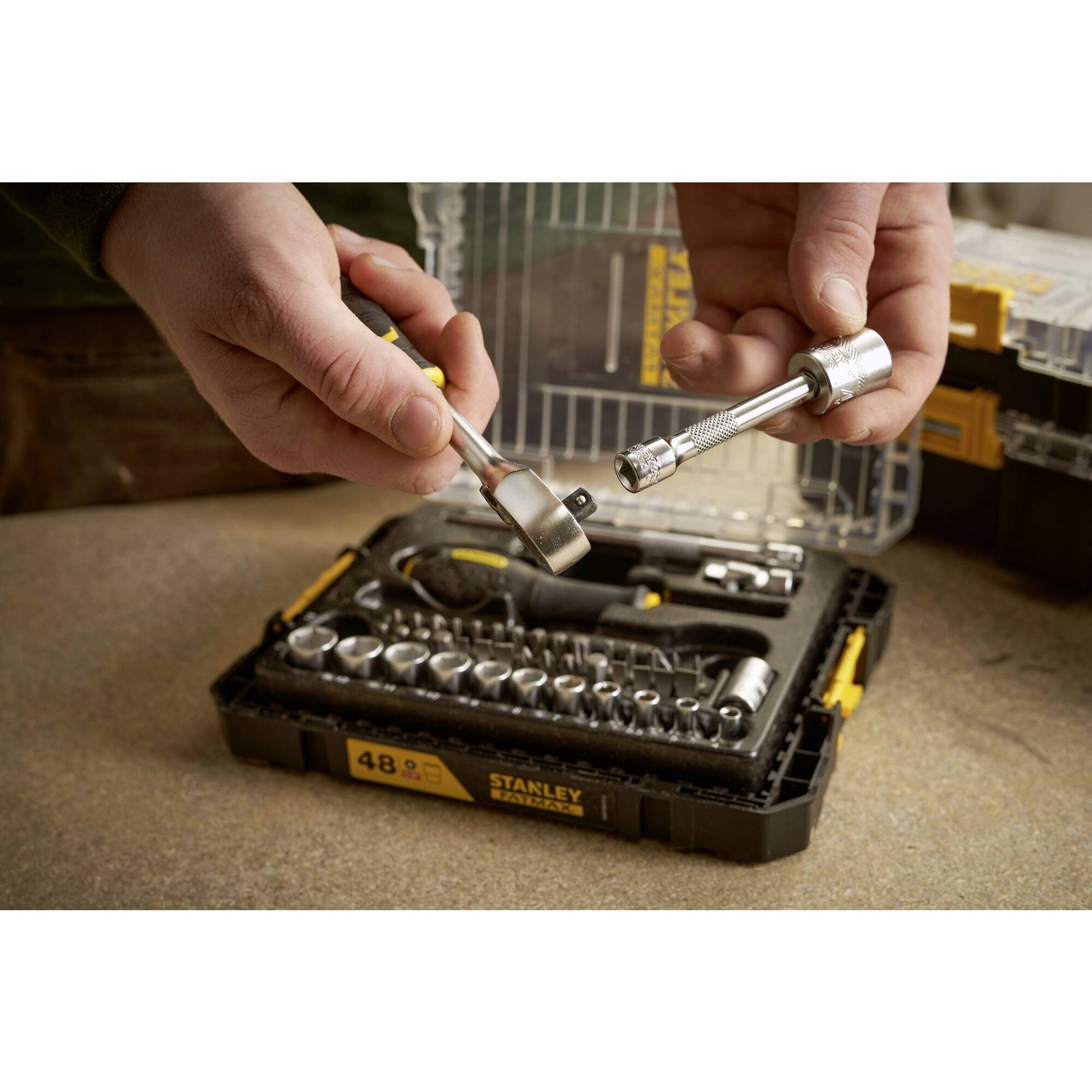 Otwarta skrzynka narzędziowa z różnymi kluczami nasadowymi. Dwie ręce mocują adapter do przedłużacza.