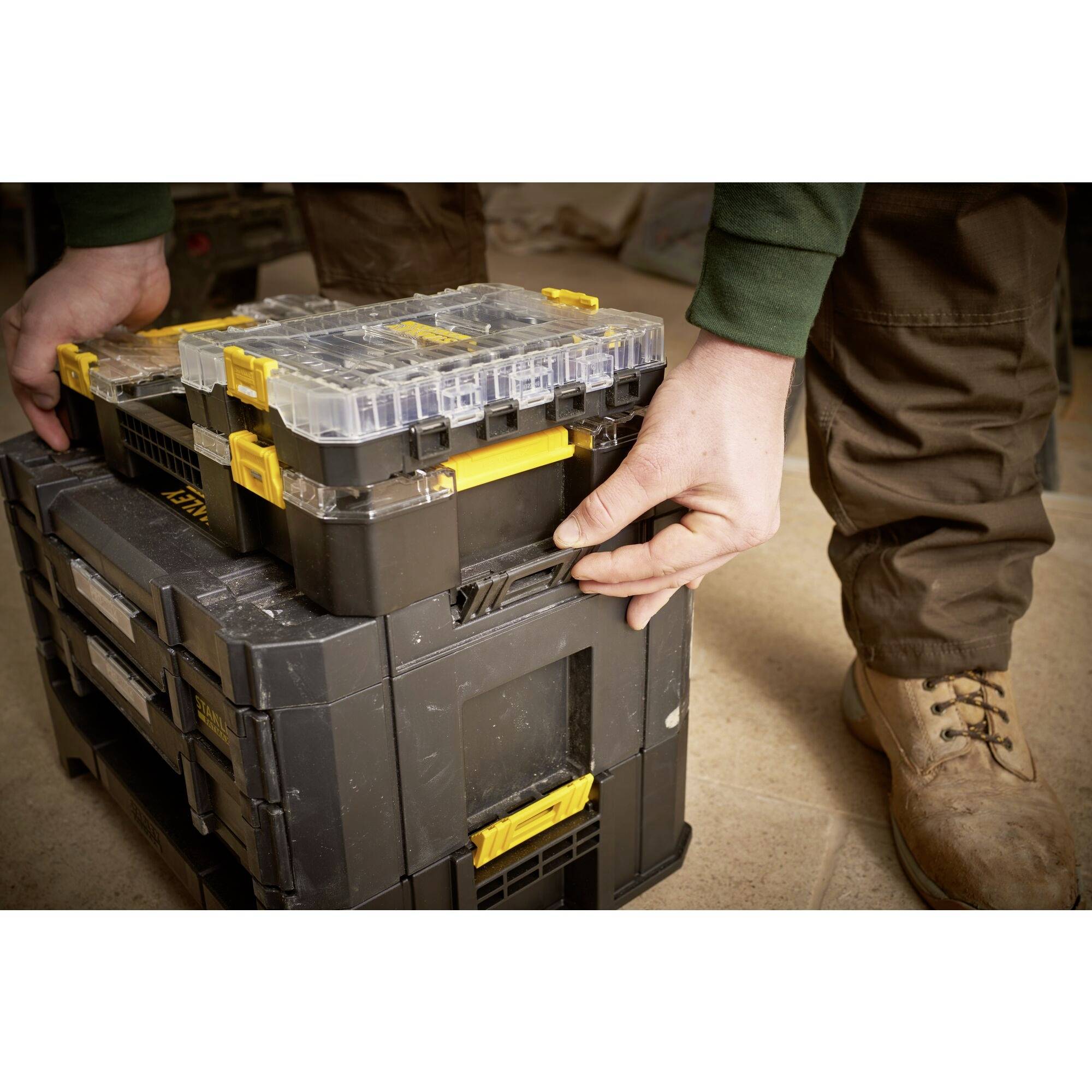 Osoba układa skrzynie z żółtymi uchwytami jedna na drugiej, prawdopodobnie podczas pracy rzemieślniczej lub budowlanej.