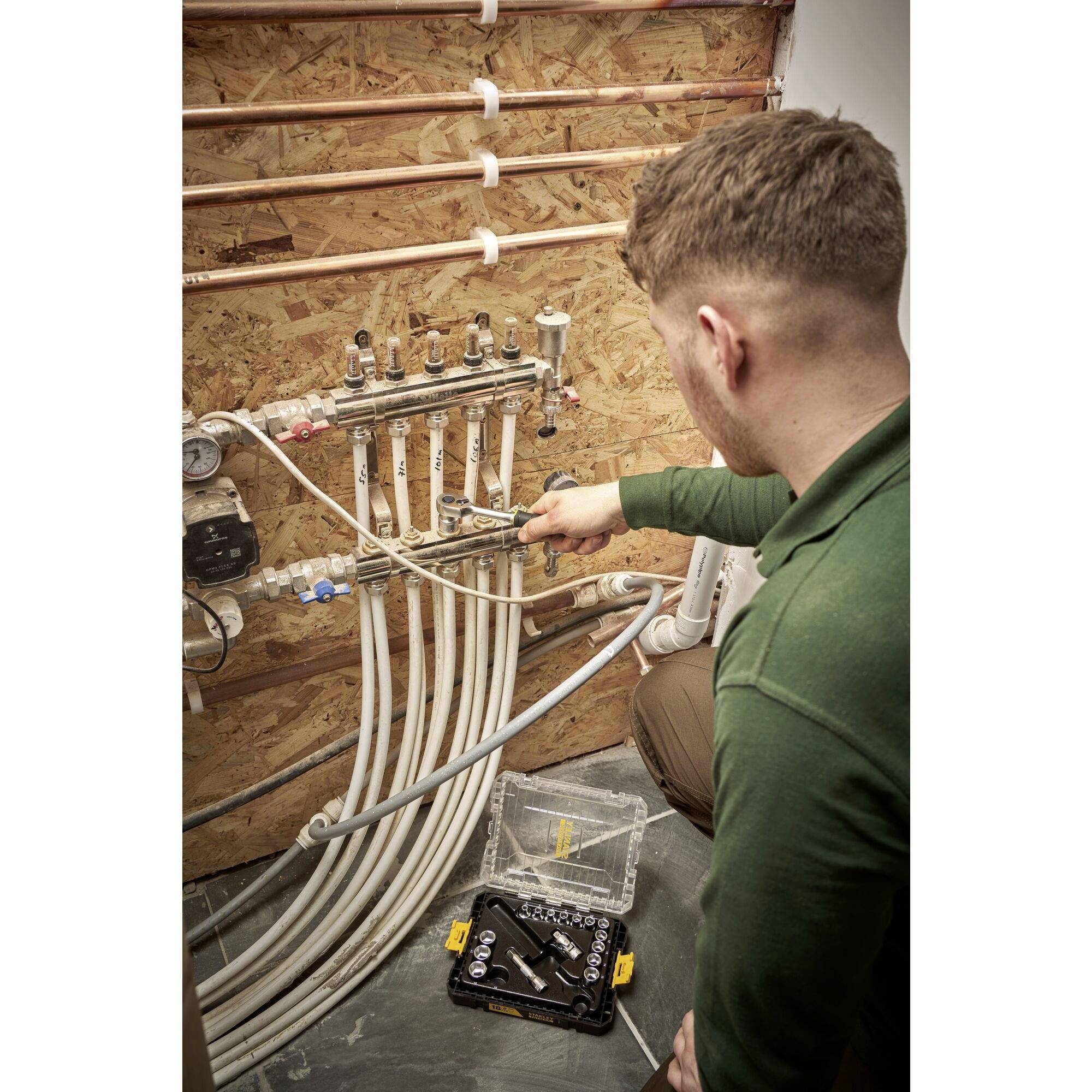 Mężczyzna pracuje przy rozdzielaczu grzewczym z wieloma rurami w pomieszczeniu technicznym. Używa narzędzia do dokonania regulacji.