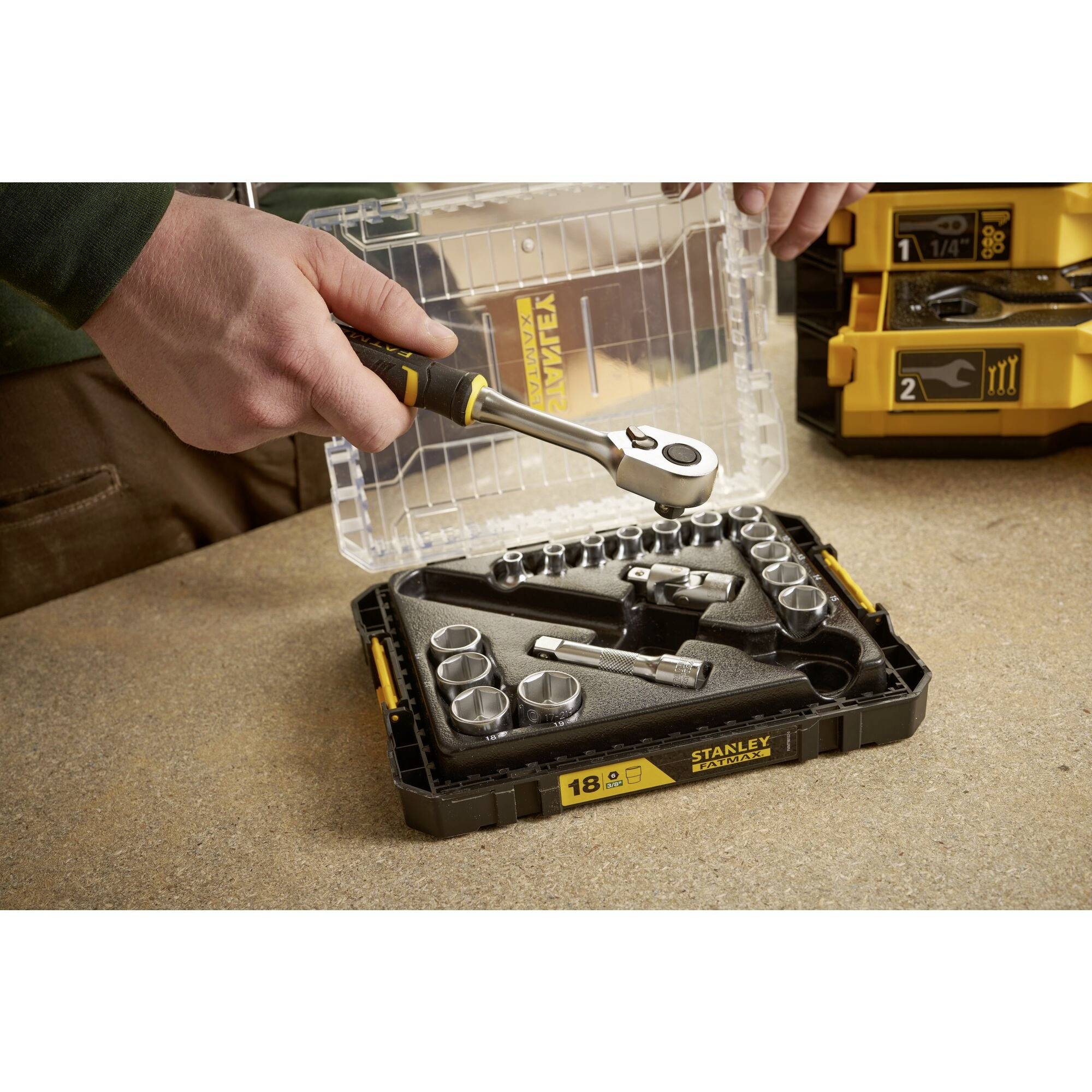 Osoba trzyma grzechotkę nad zestawem kluczy nasadowych leżącym na stole. Zestaw zawiera różne nasadki.