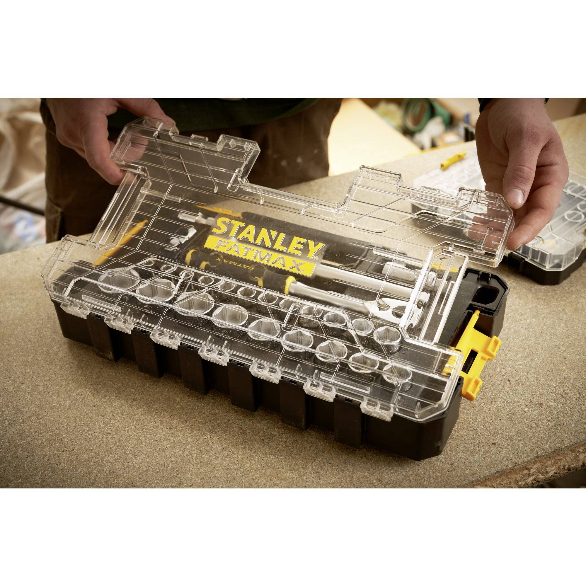 Osoba otwiera skrzynkę narzędziową z przezroczystą pokrywą, wypełnioną różnymi śrubami i narzędziami. Pudełko stoi na stole.
