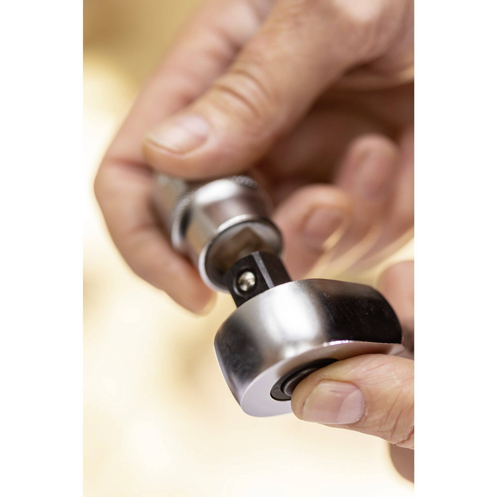 Dłoń trzyma srebrny metalowy przedmiot, prawdopodobnie narzędzie lub część, przed rozmytym tłem.