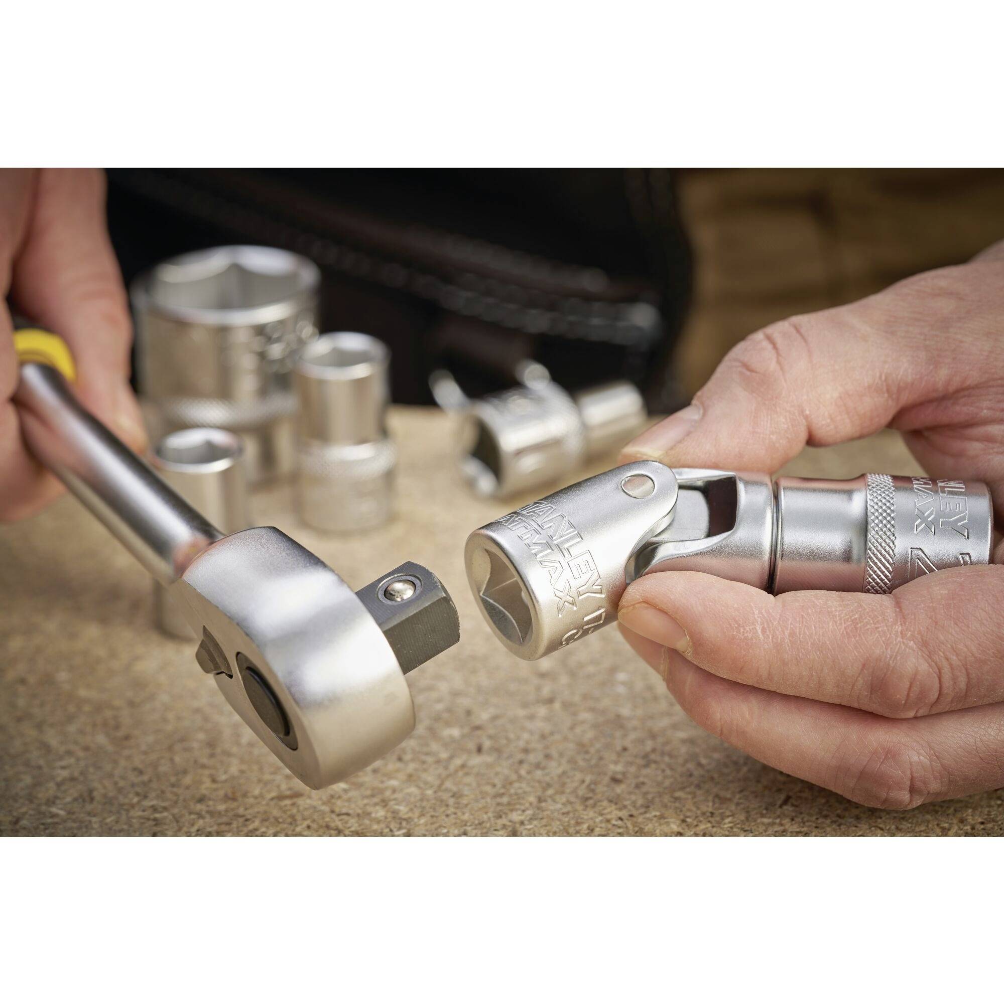 Dwie ręce montują nasadkę na klucz oczkowy z grzechotką. W tle na stole warsztatowym leżą kolejne nasadki.