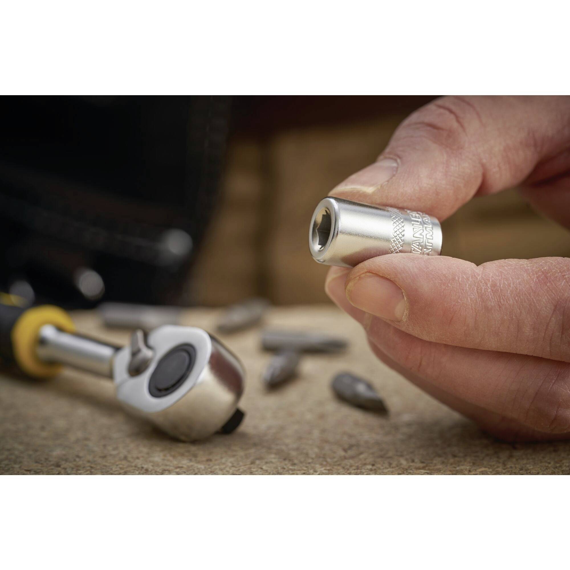 Dłoń trzyma srebrną nakrętkę, otoczoną różnymi narzędziami i elementami na drewnianym tle.