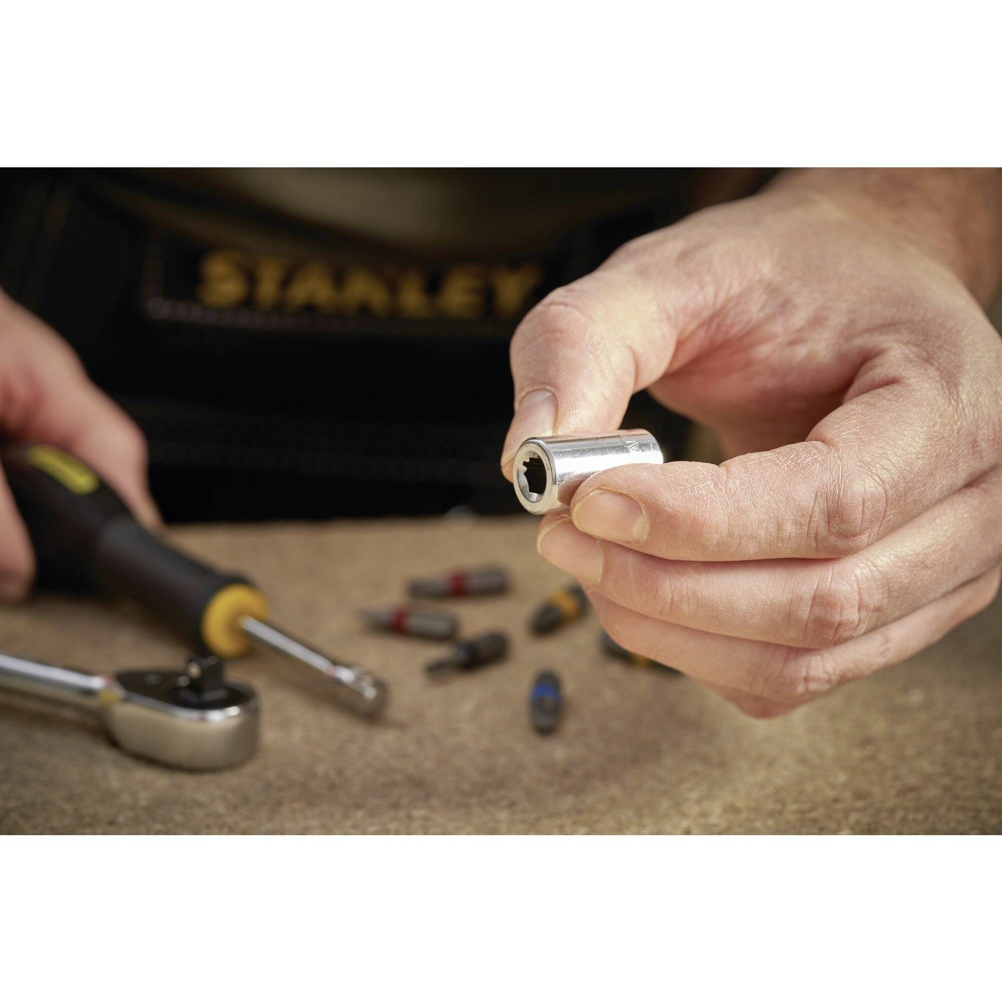 Dłoń trzyma nakrętkę przed kluczem płaskim na stole. W rozmyciu w tle widoczne są różne metalowe elementy.