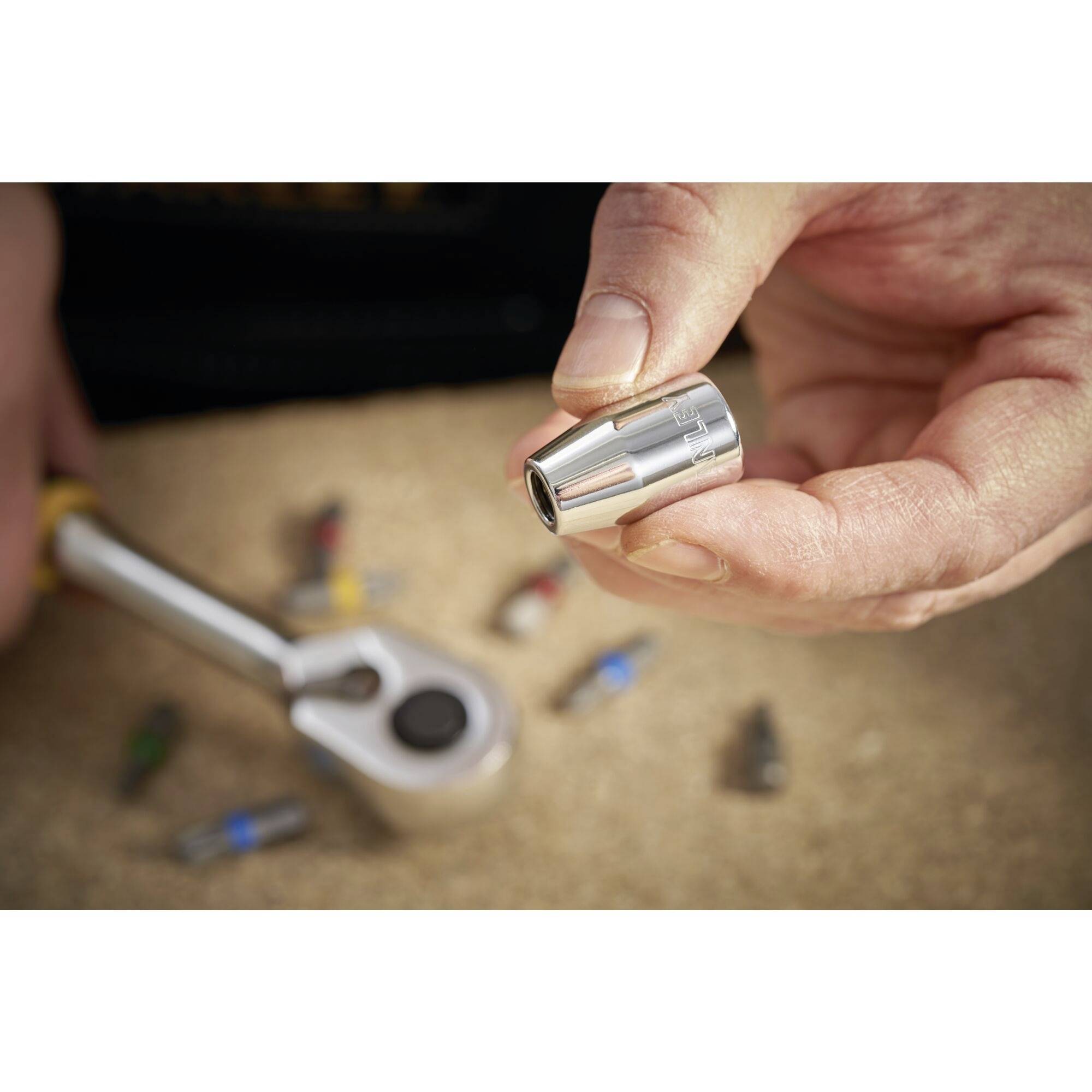 Jedna ręka trzyma srebrną wtyczkę przed stołem z rozrzuconymi narzędziami, w tym grzechotką i kilkoma końcówkami.