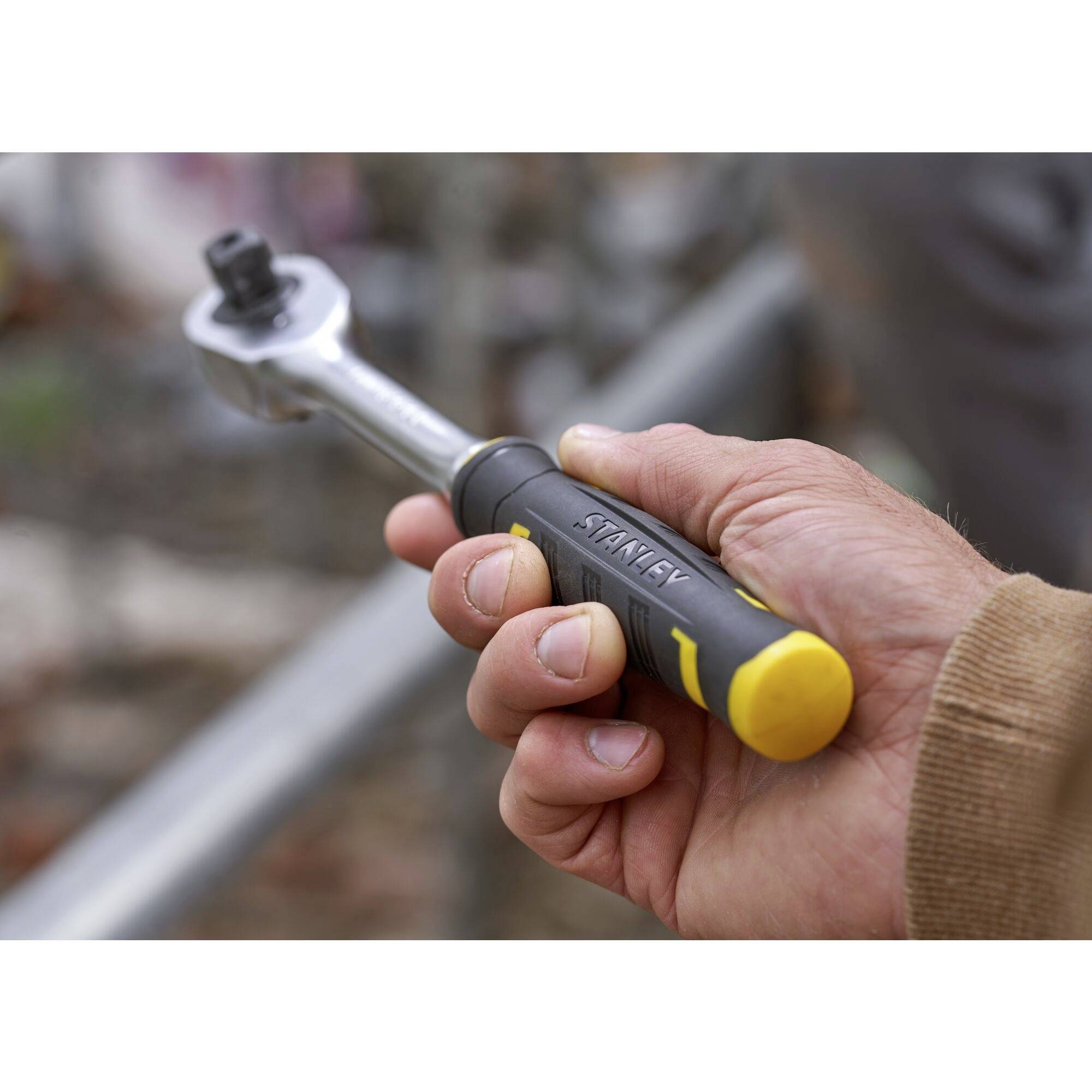 Jedna ręka trzyma klucz grzechotkowy o żółto-czarnym uchwycie, gotowy do dokręcenia śruby lub nakrętki.