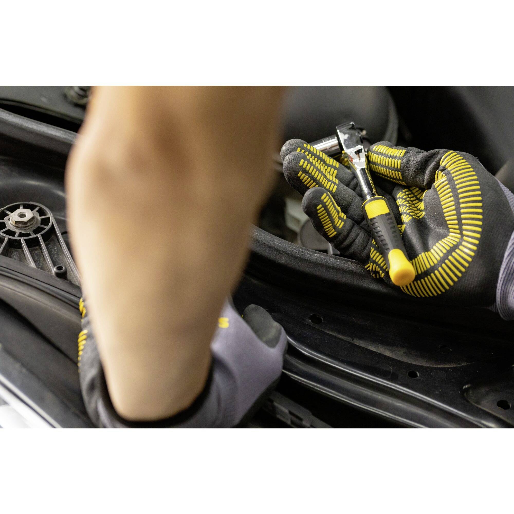 Osoba w żółtych rękawicach pracuje śrubokrętem przy części samochodowej. Zdjęcie skupia się na rękach i narzędziu.